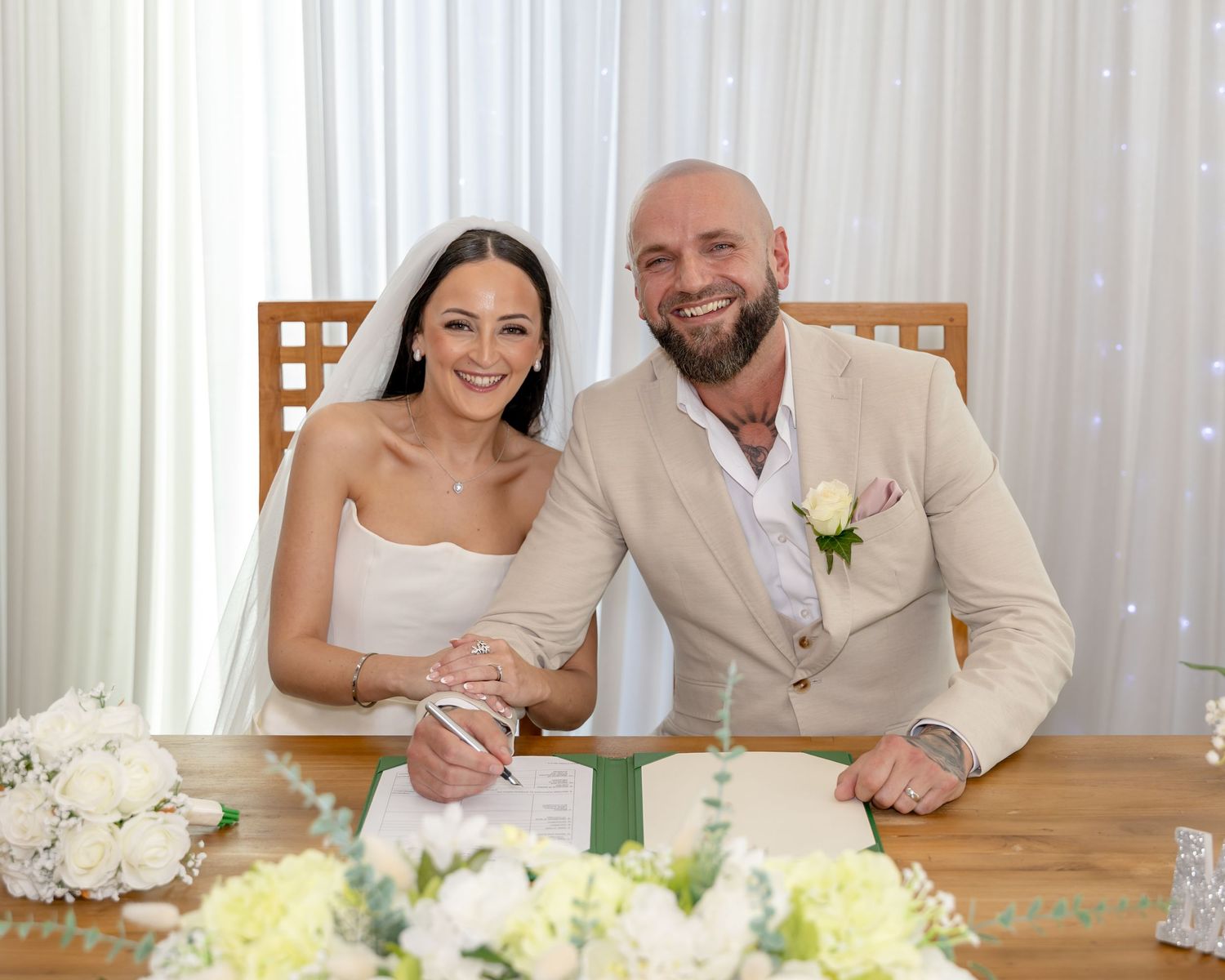 Newlyweds smile while signing wedding register surrounded by white floral arrangements.