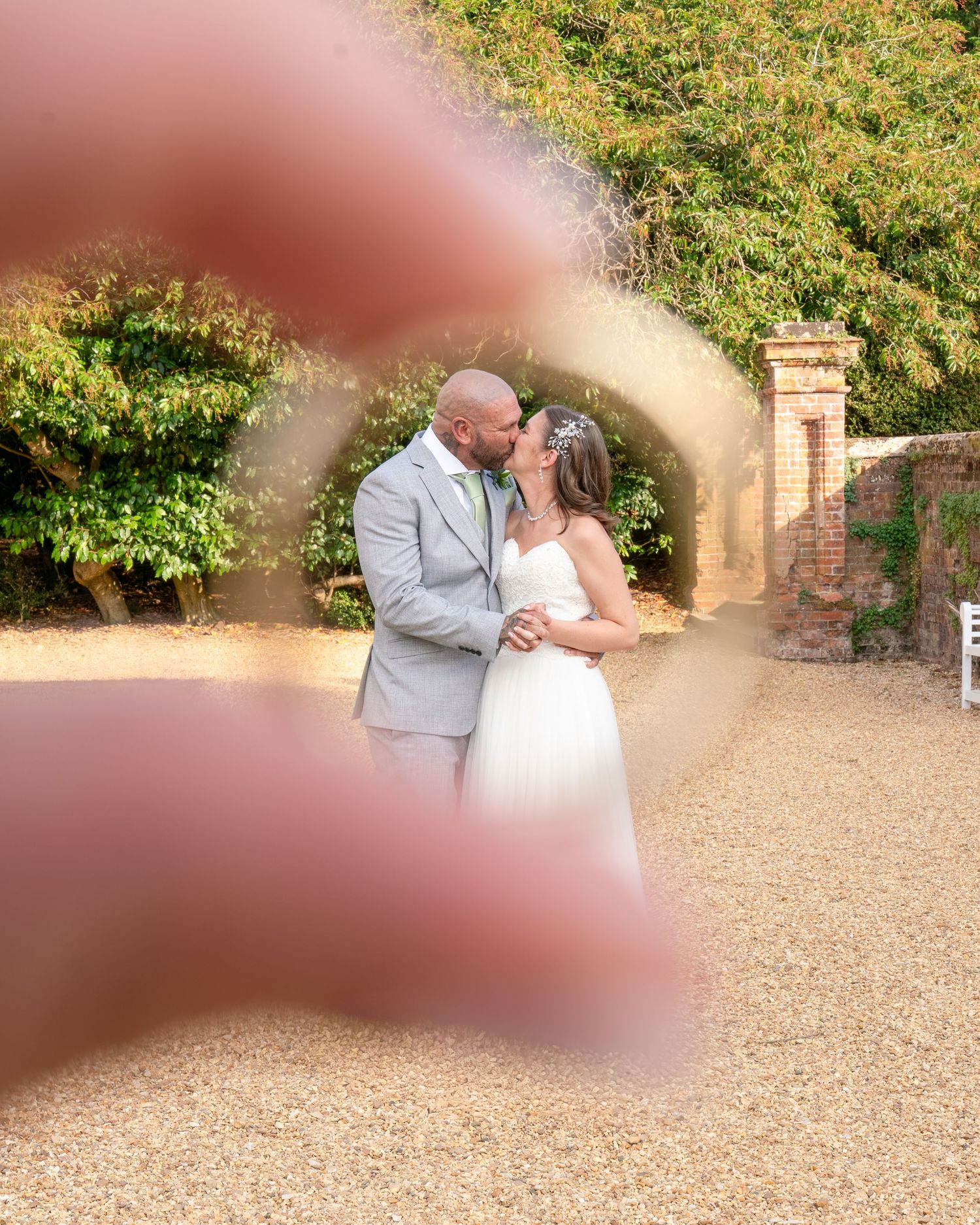 Bride and groom kissing framed by a ring gesture at an outdoor wedding venue with brick archway and lush greenery.