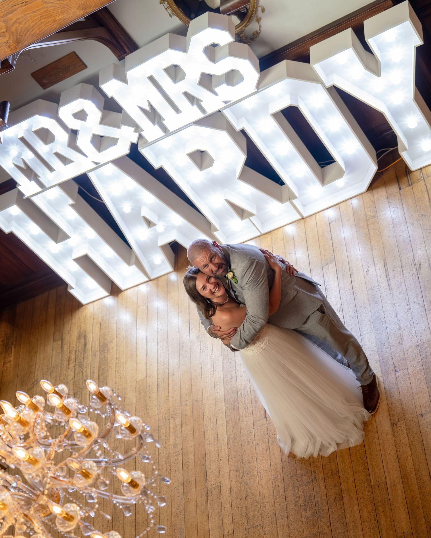 Newlyweds Mr. & Mrs. Hardy embrace in front of illuminated marquee letters at their wedding reception.