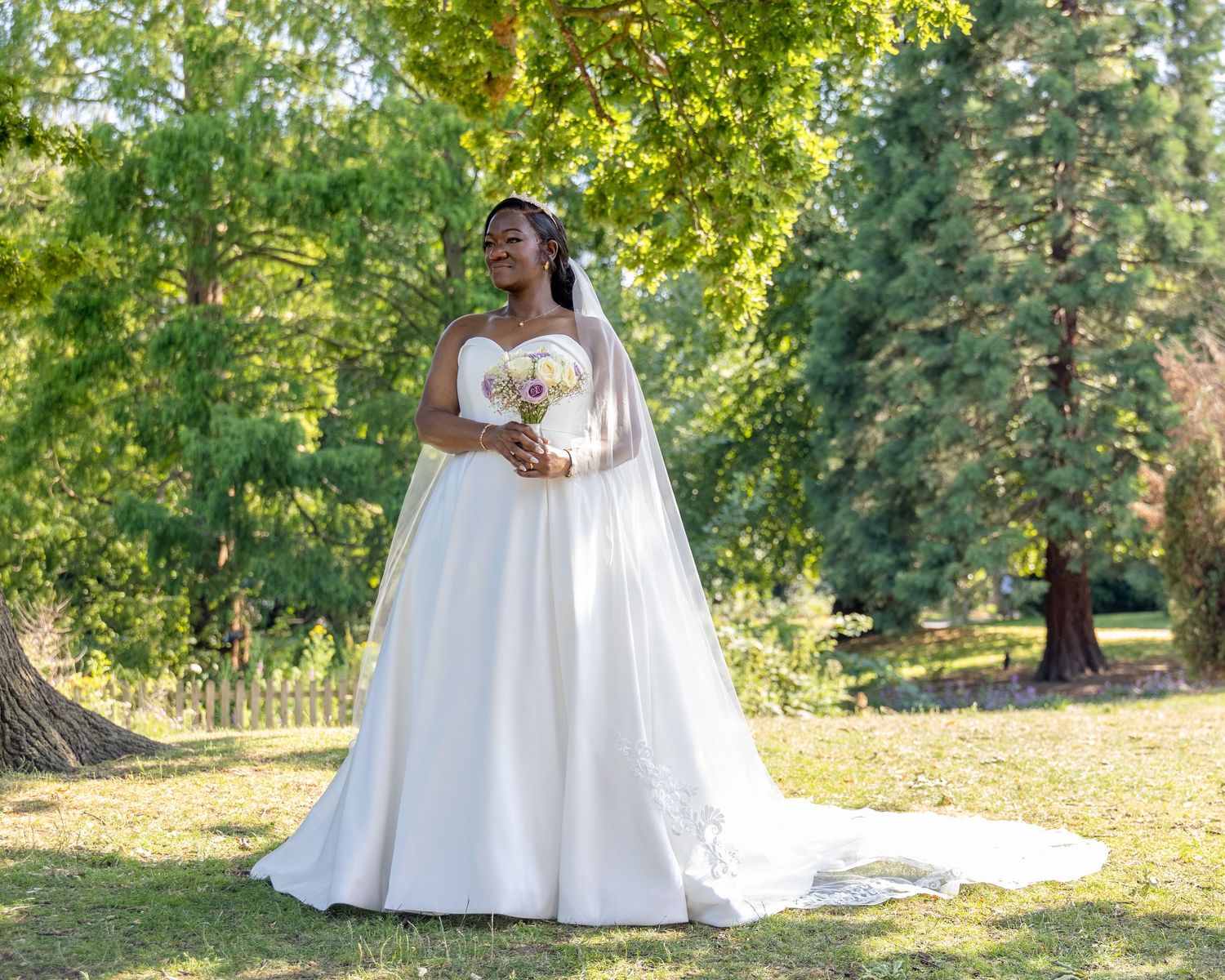 Bride in white ball gown wedding dress holding bouquet, standing in lush green park setting.