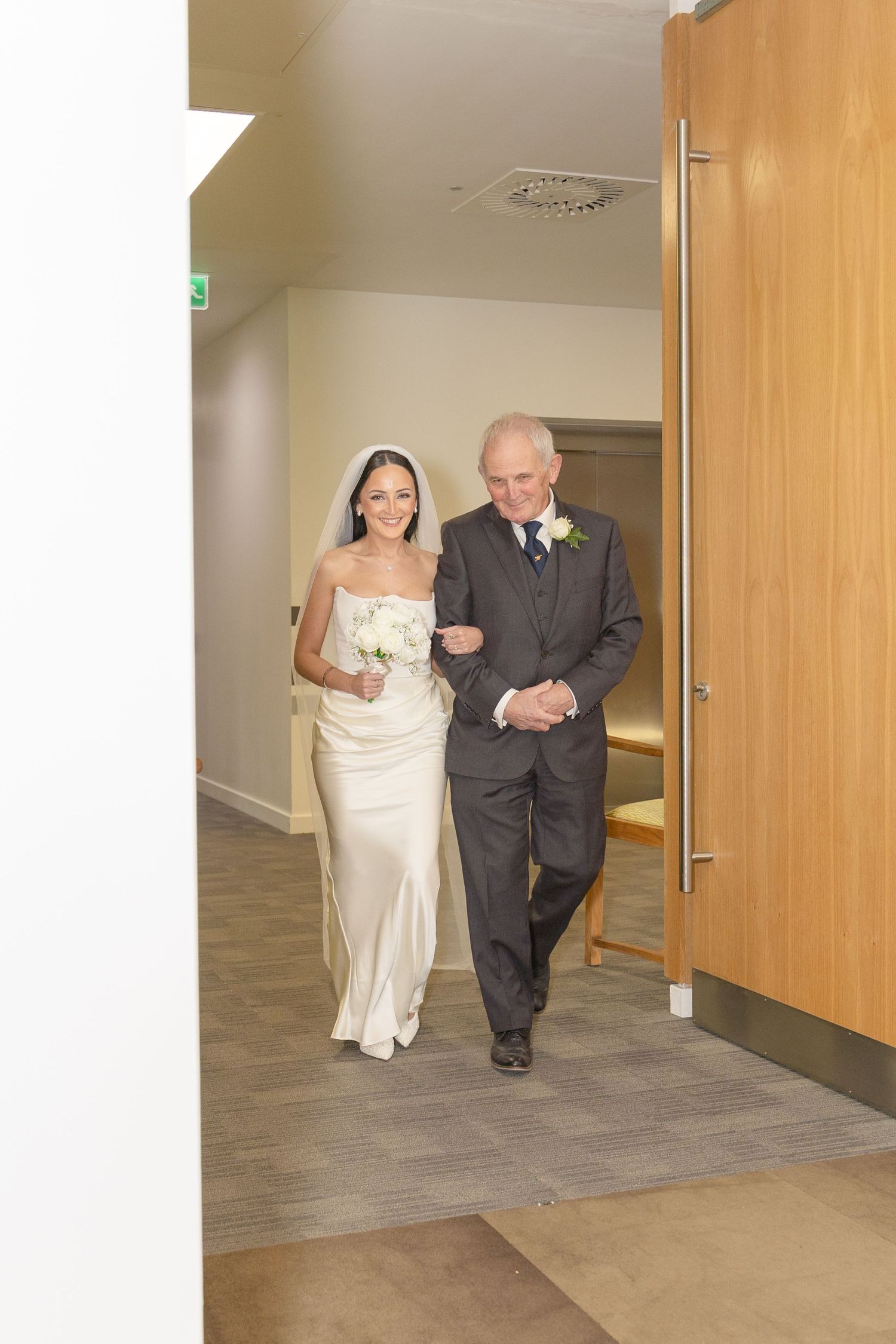 Bride wearing white veil and satin gown walks with suited older man through a bright hallway toward ceremony.