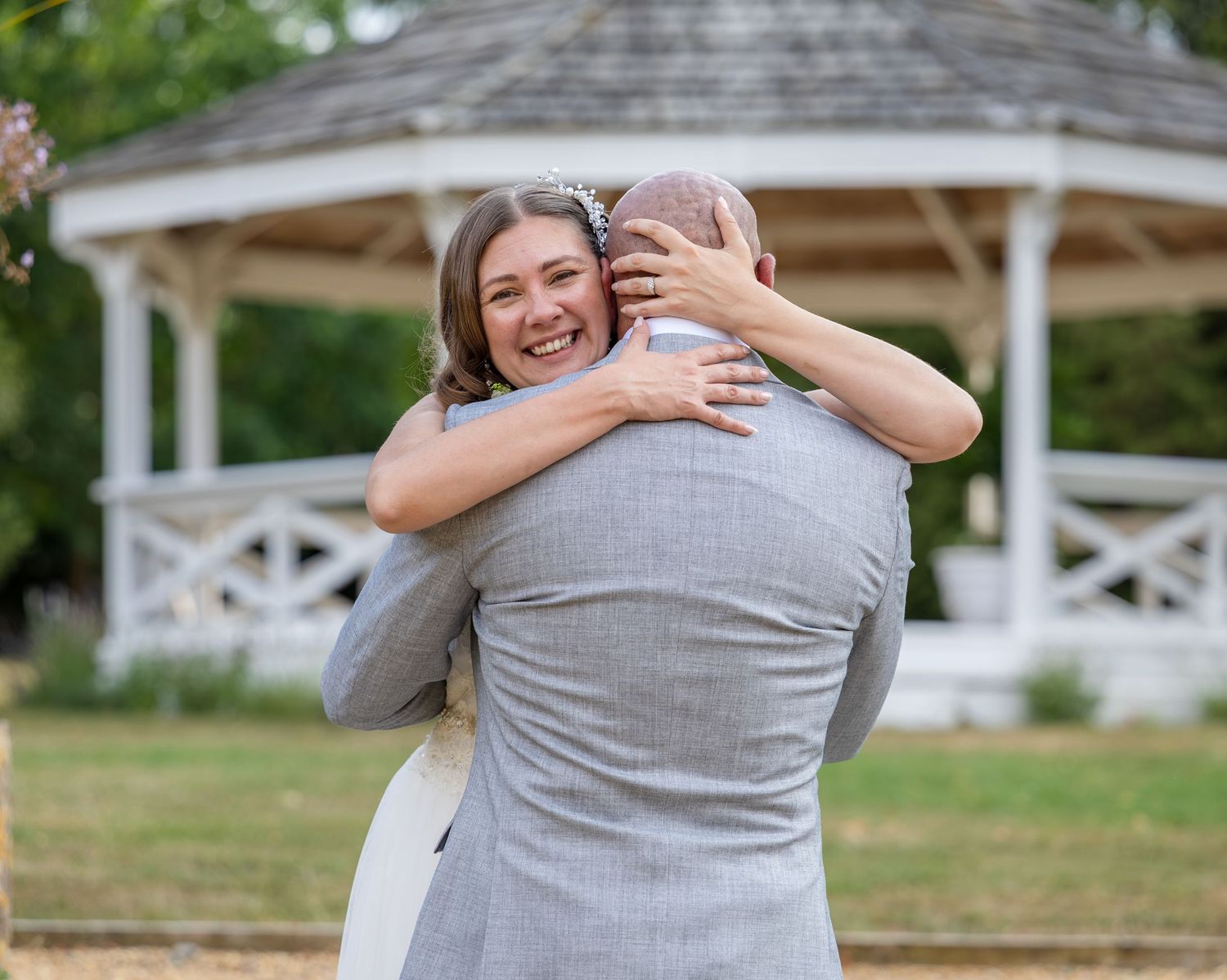 Bride and groom sharing a warm embrace near a white gazebo during an outdoor wedding ceremony.