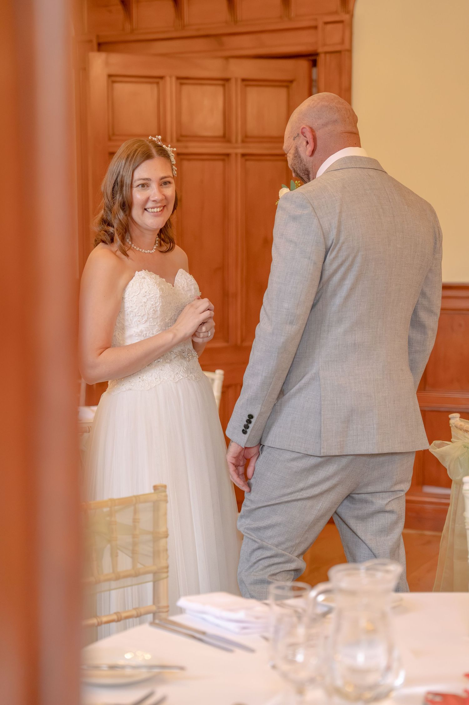 Bride in white gown smiles at bald groom in grey suit during wedding reception in elegant wood-paneled venue.
