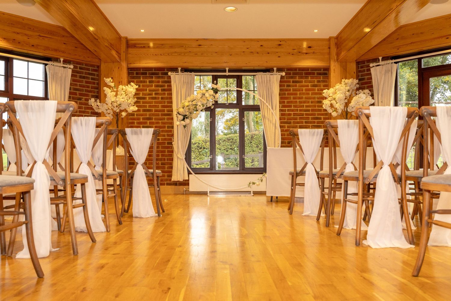 Elegant wedding ceremony setup with wooden chairs, white draping, and floral arrangements in a rustic venue.