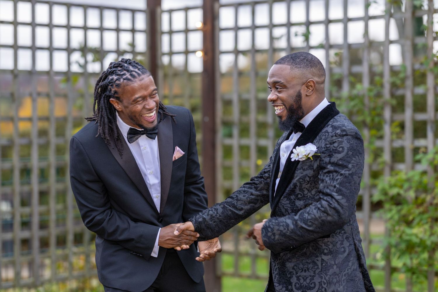 Two men in formal tuxedos laugh and shake hands at a wedding celebration outdoors.
