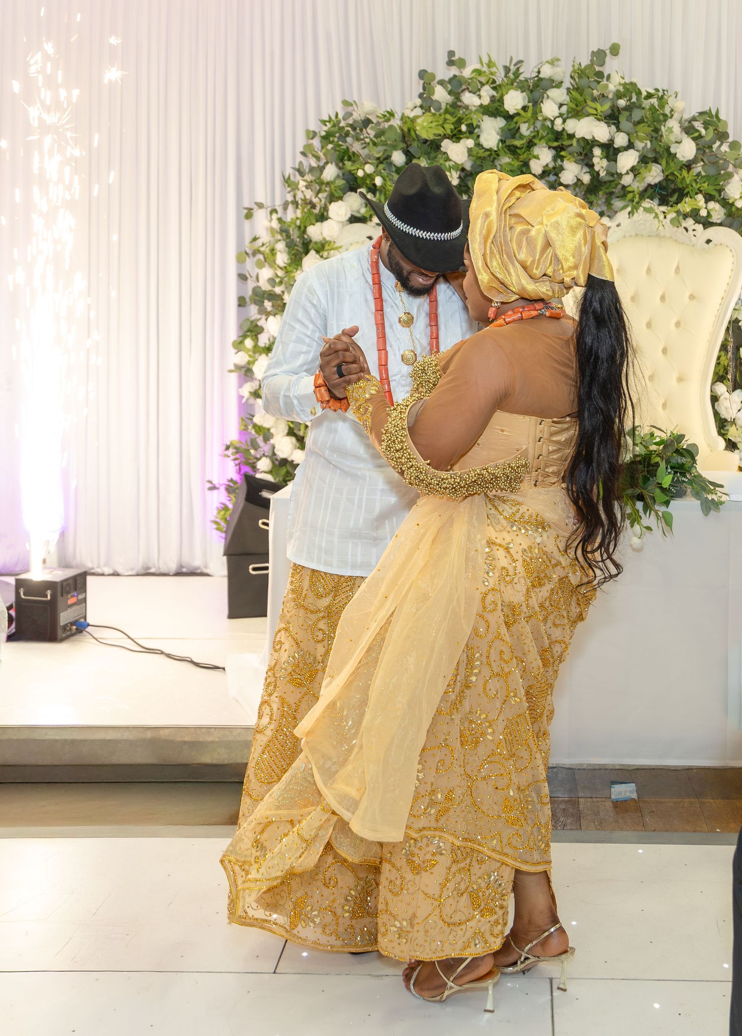 Nigerian bride and groom sharing intimate moment in gold aso-oke and white traditional attire before floral wedding backdrop.