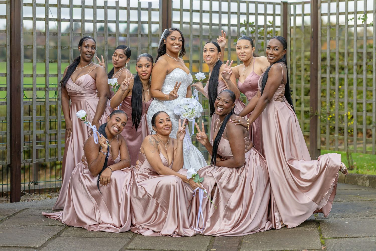 Bridesmaids in matching pink satin dresses pose elegantly against a garden trellis.