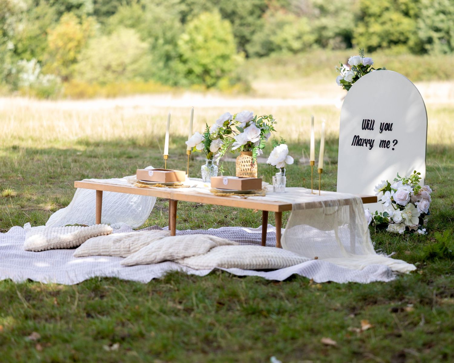 Elegant outdoor proposal setup with white flowers, candles, picnic blanket and Will You Marry Me sign.