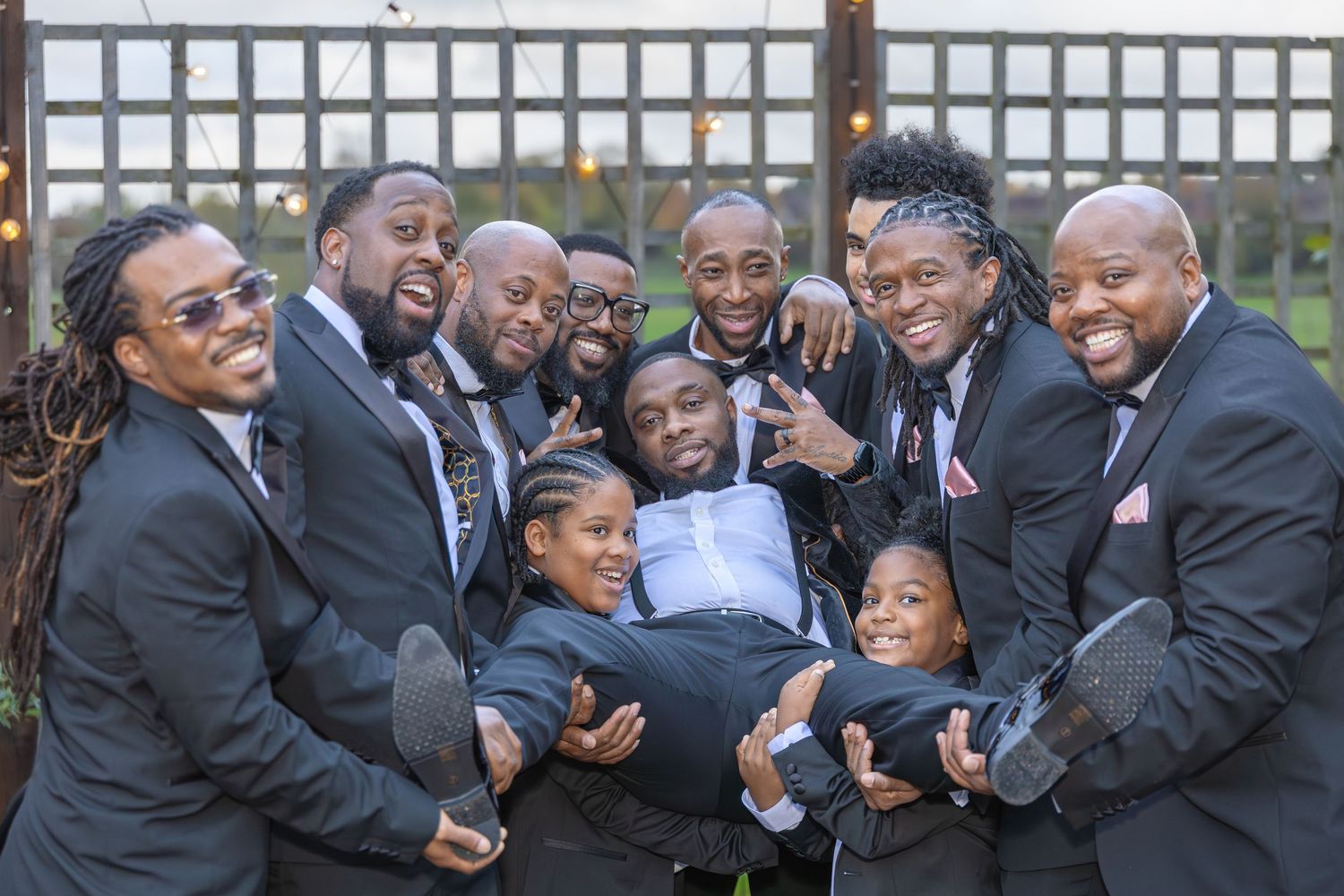 Groomsmen in black suits joyfully lift the groom while two young boys join the fun wedding party photo.