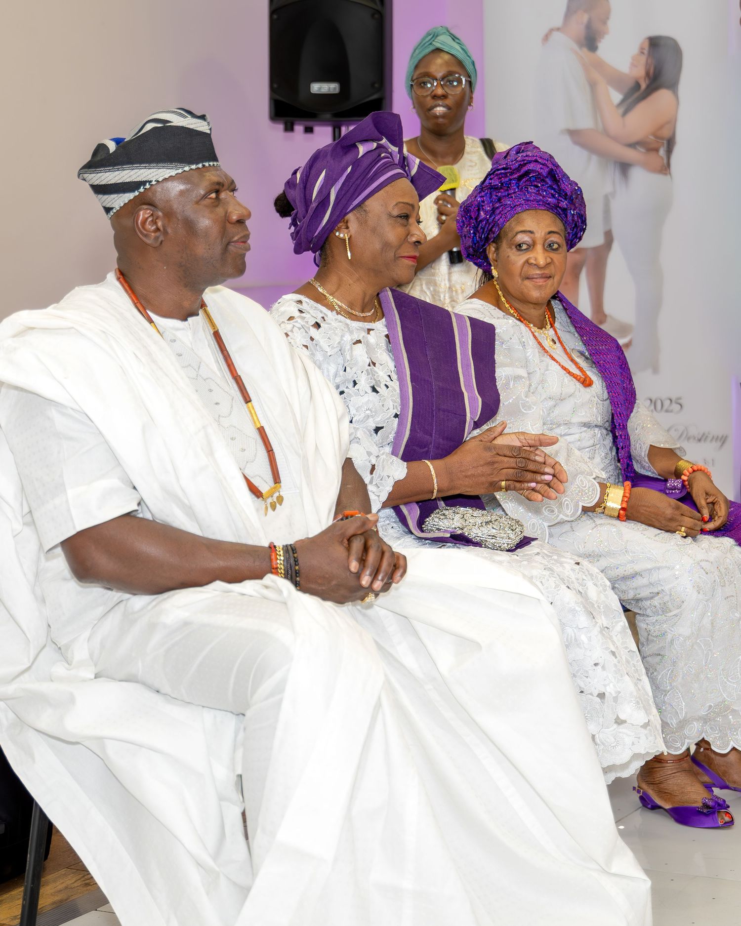 Three guests in traditional Nigerian attire of white and purple aso-oke sit together at a cultural wedding ceremony.