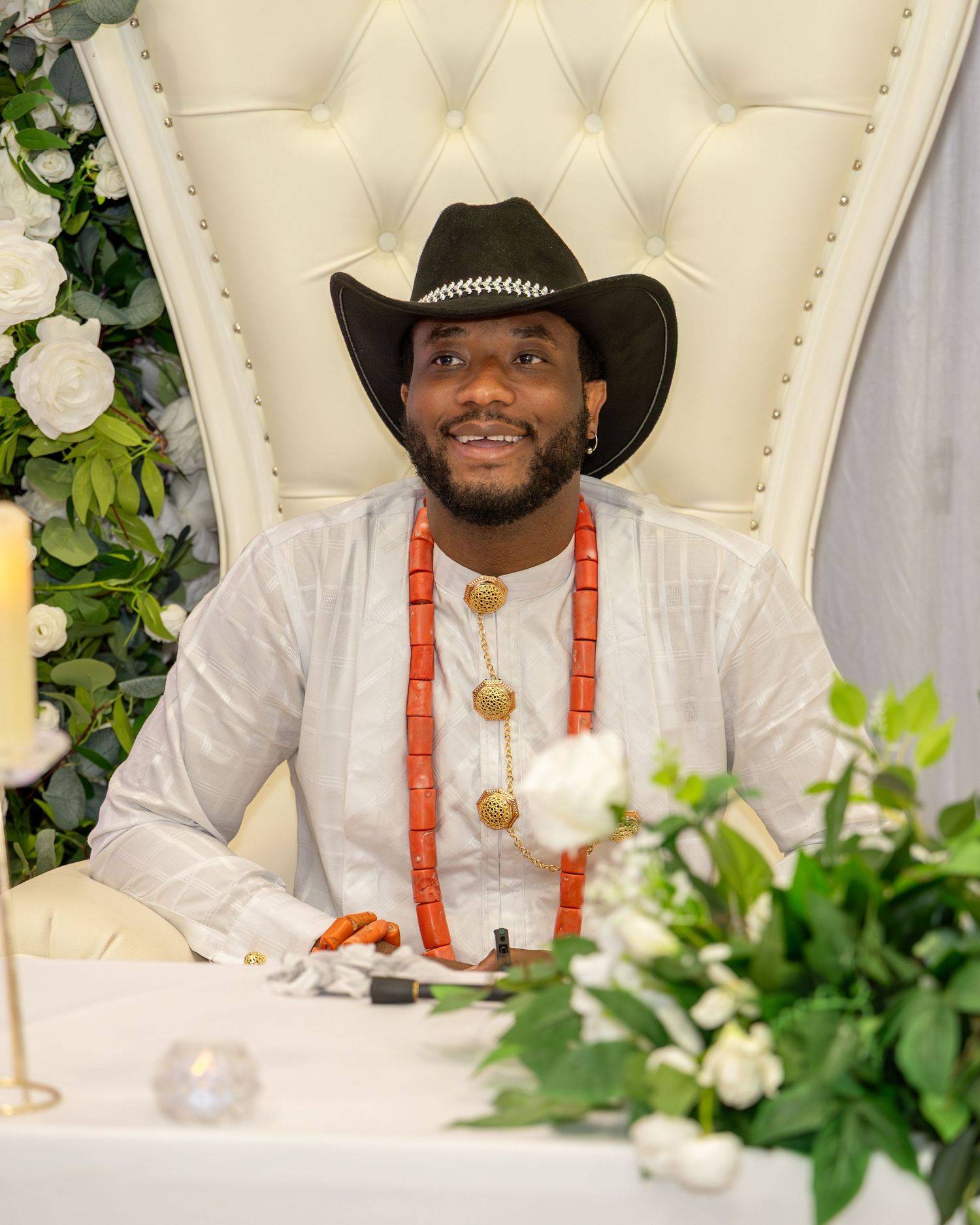 Groom in white Nigerian traditional outfit with coral beads and black cowboy hat seated at decorated wedding throne chair.