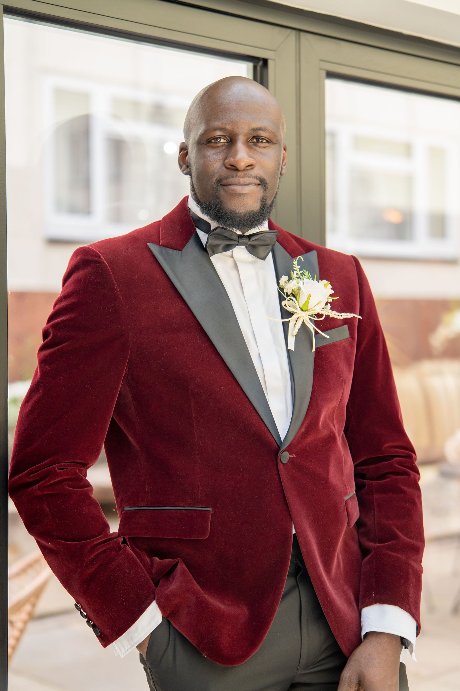 A well-dressed Black man in a burgundy velvet tuxedo jacket with bow tie and boutonniere poses confidently outdoors.