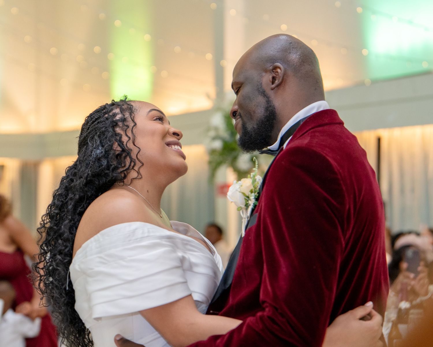 Couple sharing first dance at wedding reception, bride in white off-shoulder gown, groom in red velvet suit.