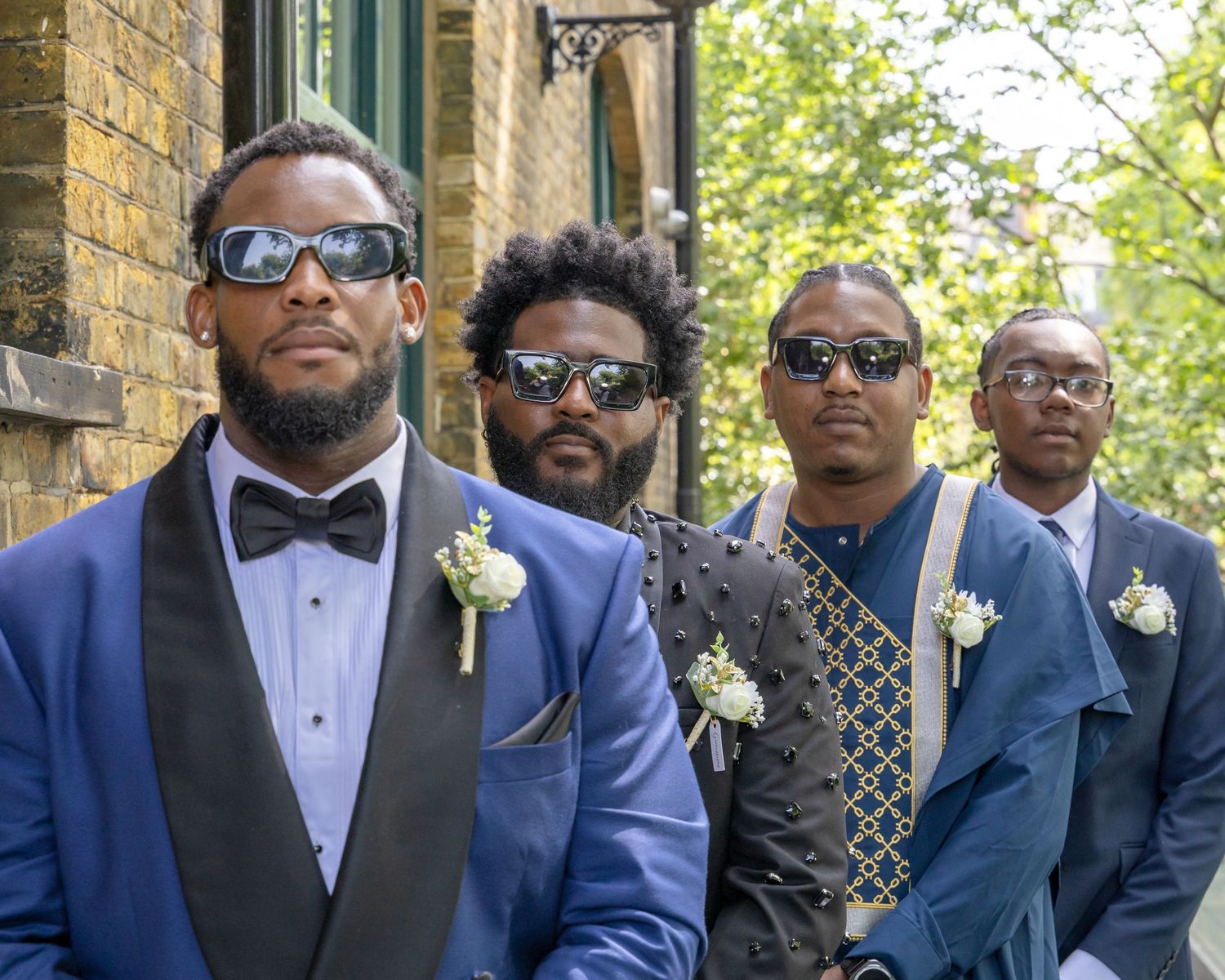 Four men in blue suits with boutonnieres and sunglasses pose outdoors at a formal wedding event.