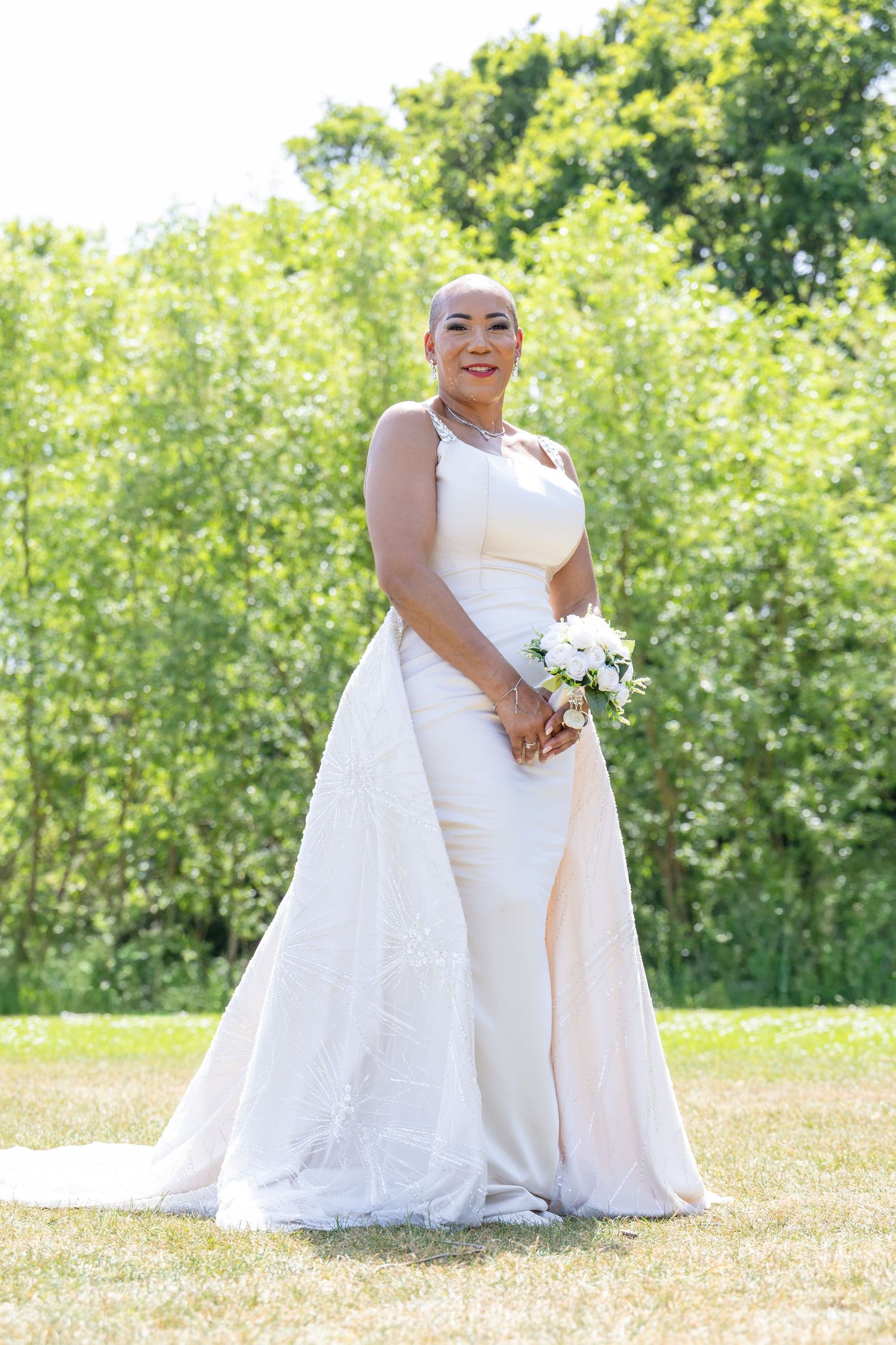 Smiling bride in white fitted gown with train holds bouquet outdoors in lush green park on sunny day.