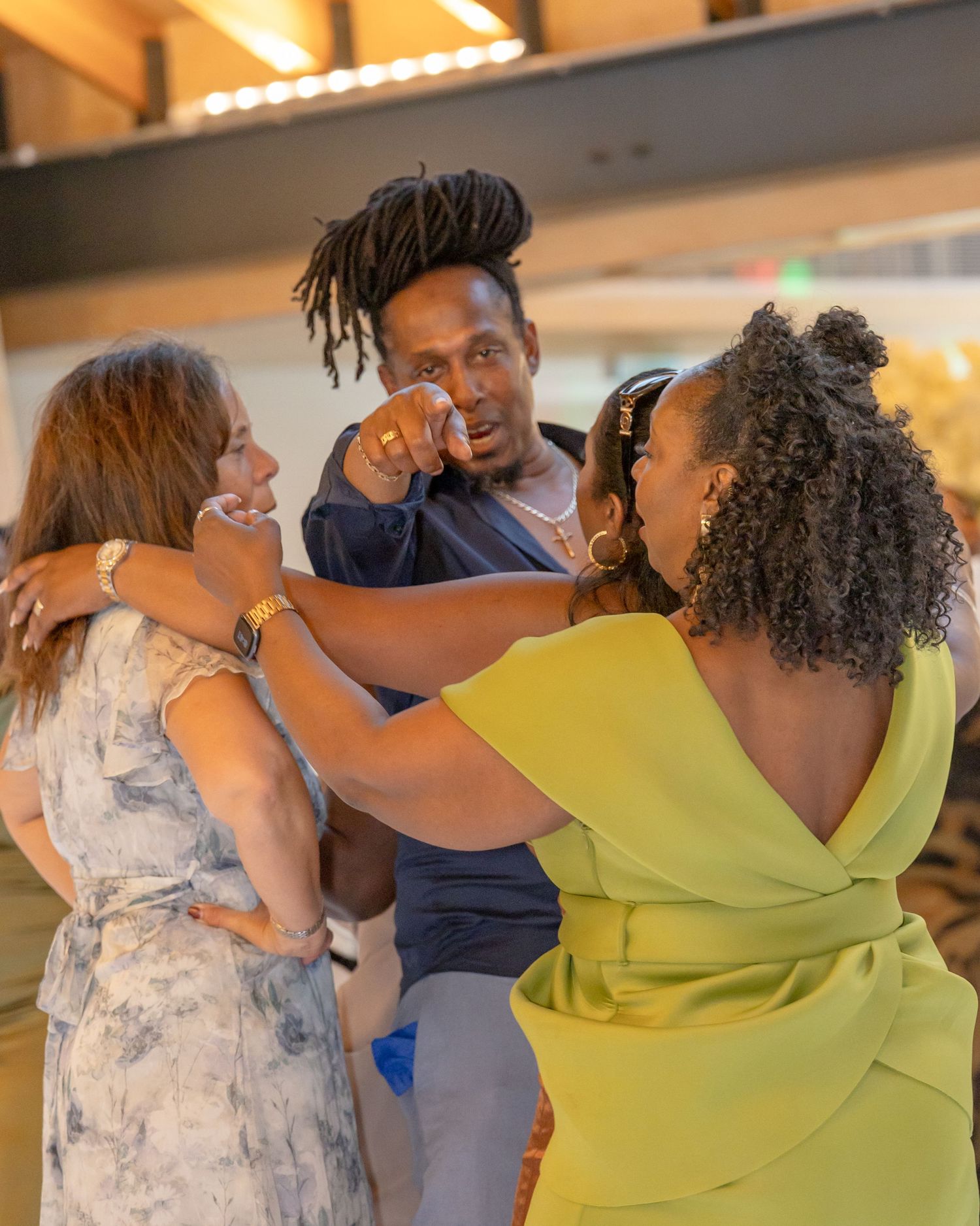 Three people sharing a joyful emotional embrace at an indoor event, with a man wiping tears while two women smile.