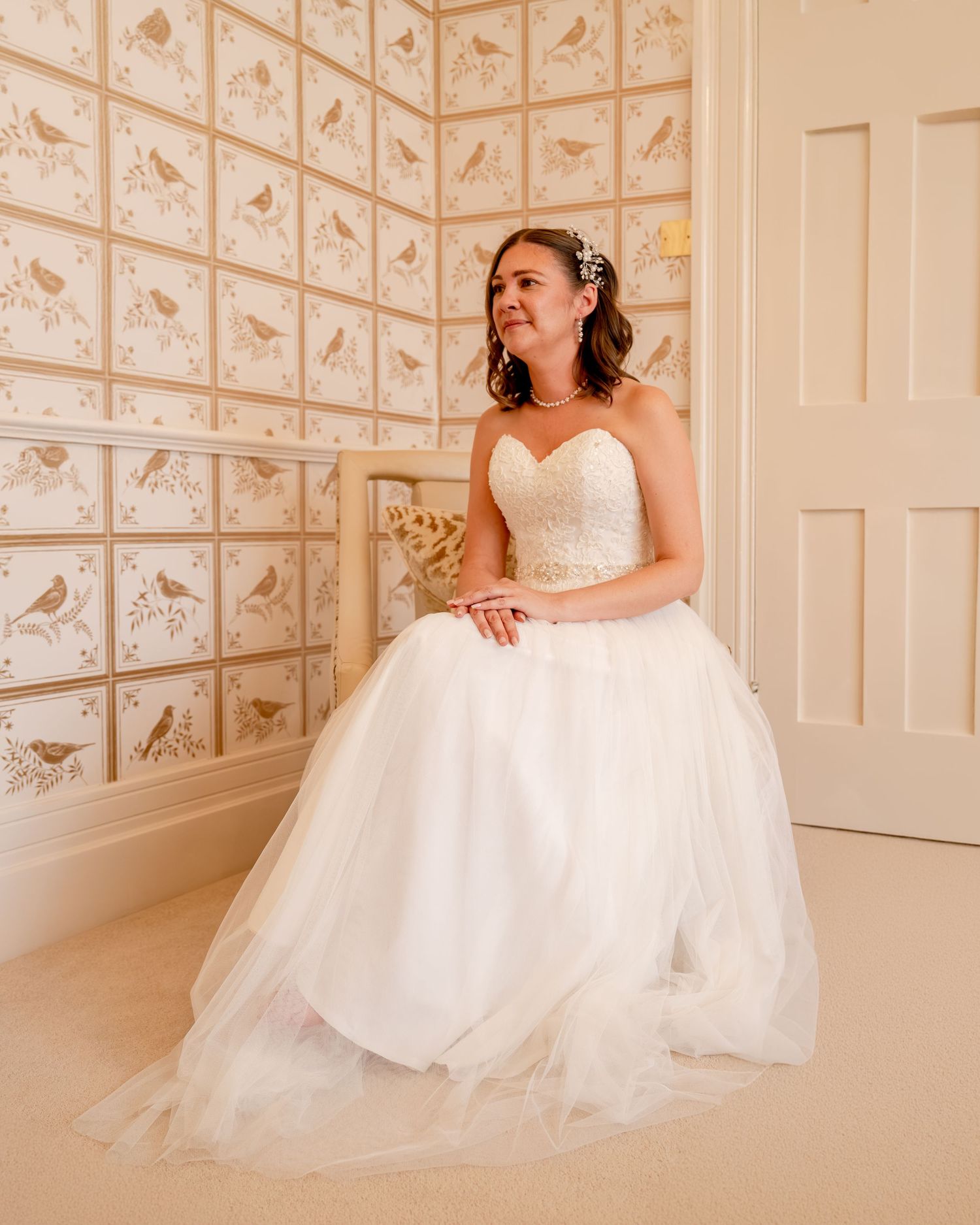 Bride in strapless white tulle ball gown sits gracefully in a room with elegant bird-patterned wallpaper.