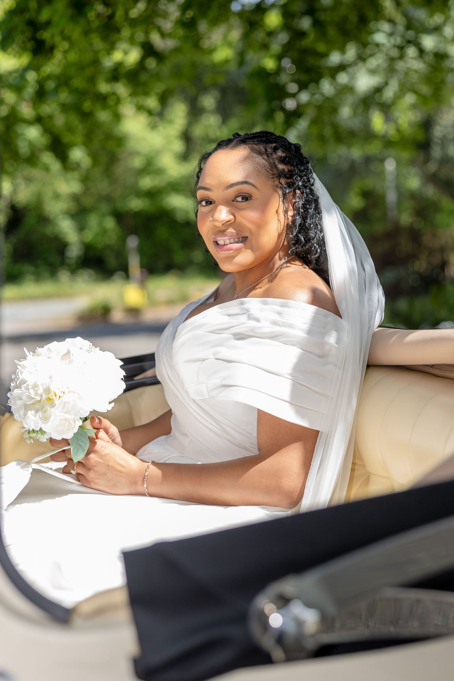 Smiling bride in white off-shoulder gown holding white bouquet, seated in elegant carriage surrounded by lush green trees.