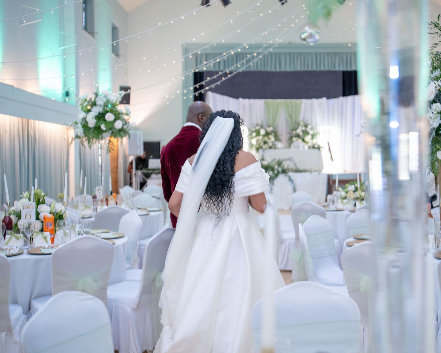 Bride and groom walking through an elegantly decorated white wedding reception hall with floral arrangements.