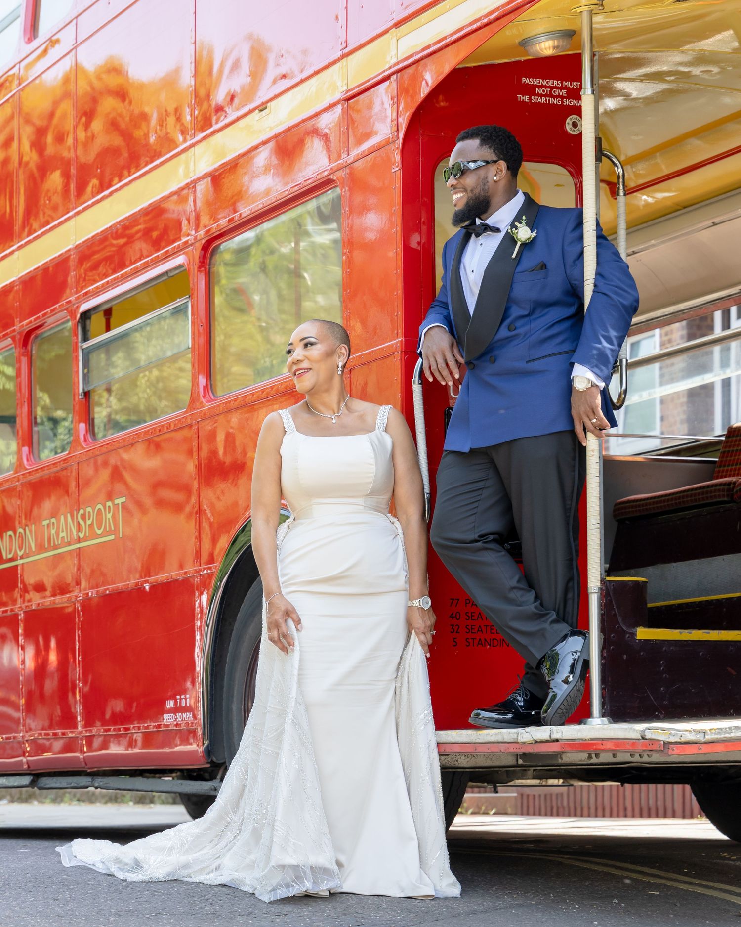 Bride in white gown and groom in blue suit pose by a red London Transport double-decker bus on their wedding day.