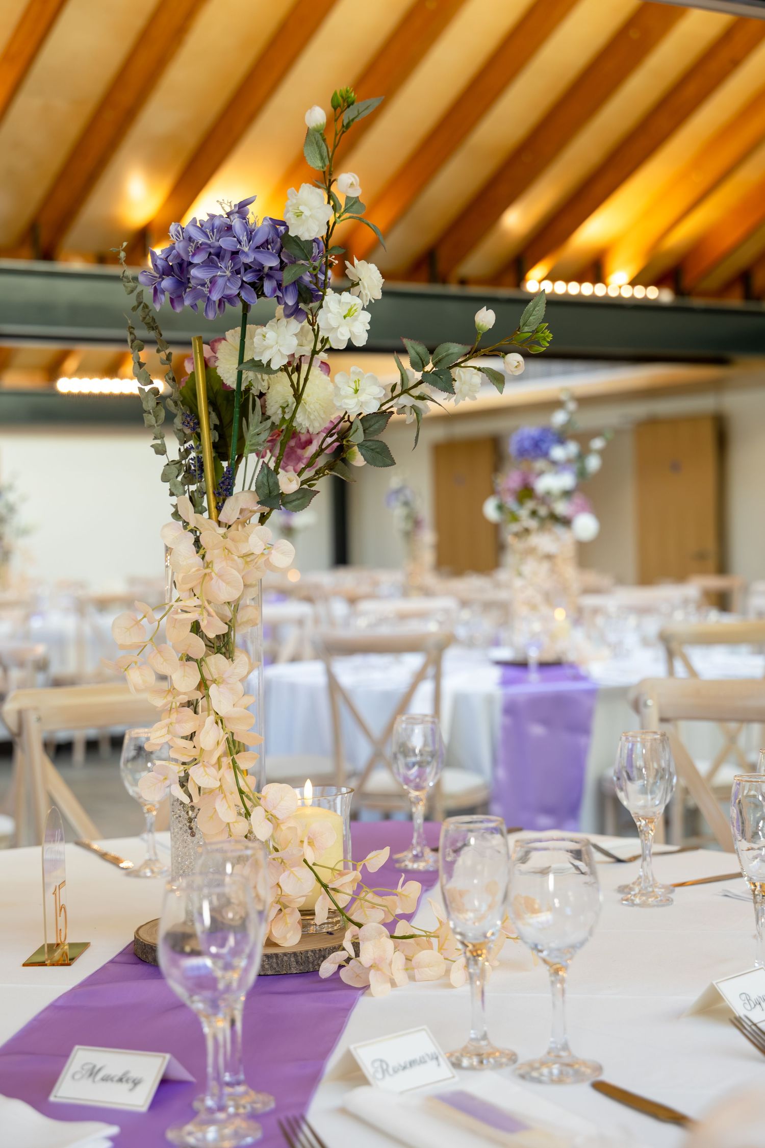 Elegant wedding reception table with purple runners, tall floral centerpiece featuring hydrangeas and white orchids in a rustic venue.