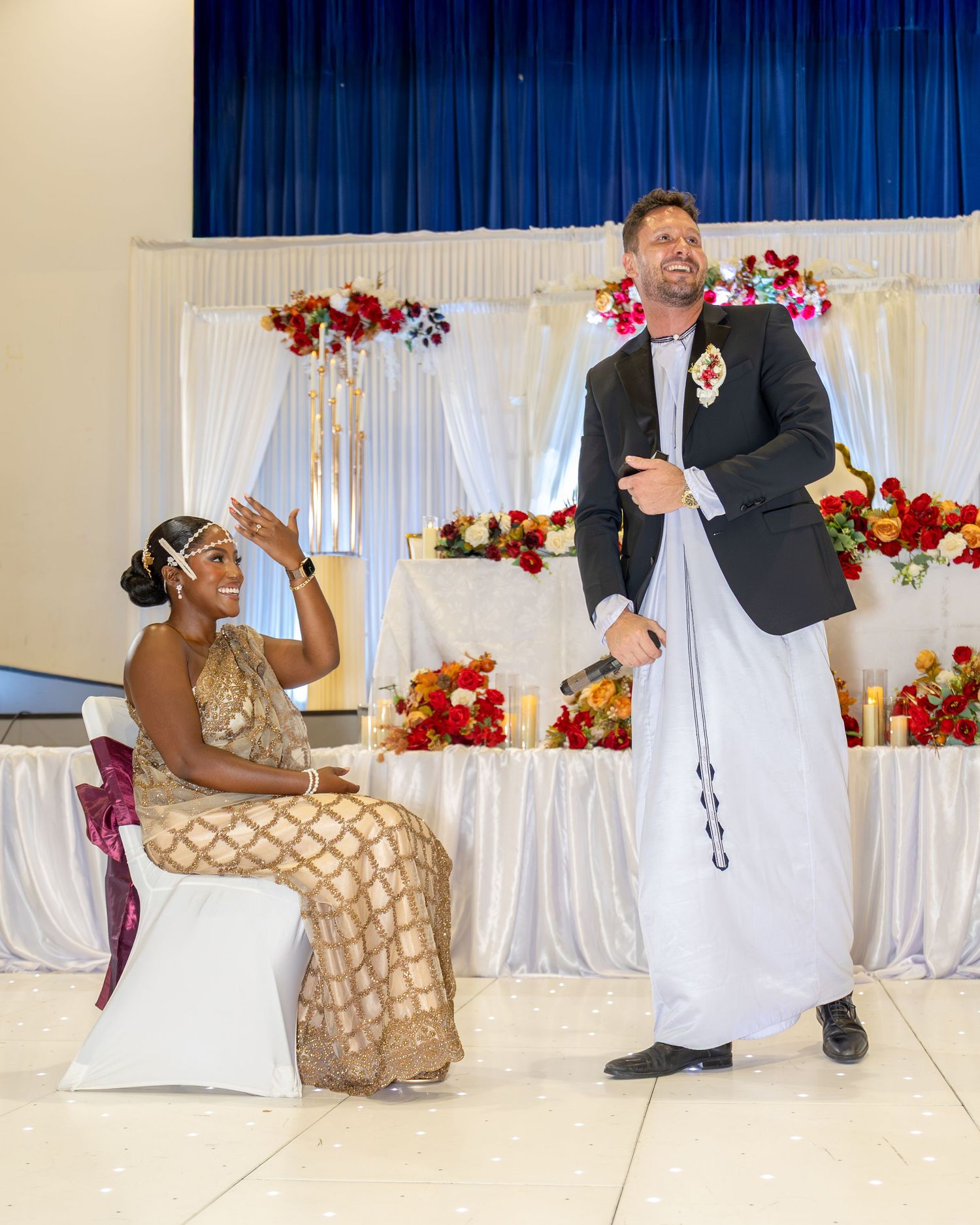 Bride in gold sequin dress sits while groom in white traditional attire stands beside her at wedding reception.