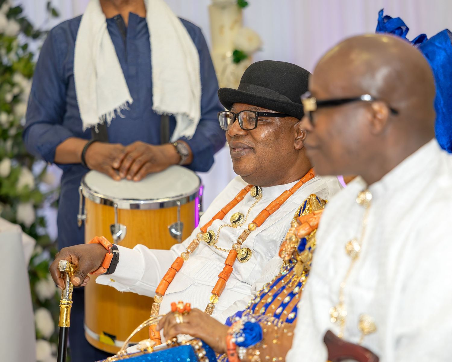 Nigerian elder in traditional attire with coral beads playing drums at a cultural wedding ceremony.