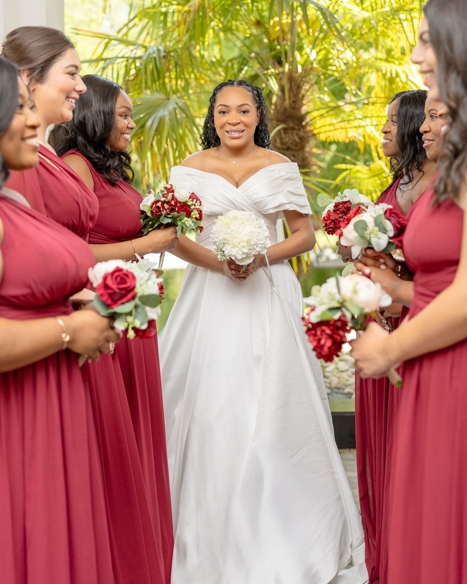 Bride in white off-shoulder gown smiles surrounded by bridesmaids in red dresses holding rose bouquets outdoors.