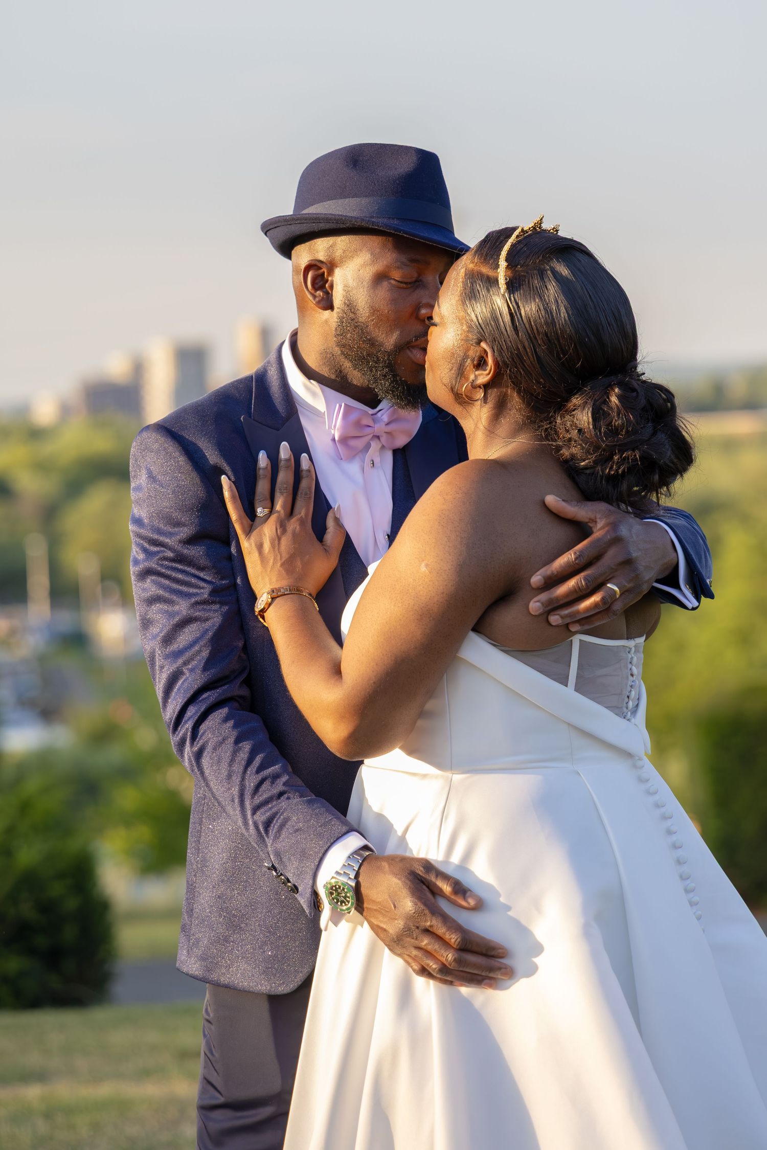 A groom in a blue suit and pink bow tie kisses his bride in a white dress outdoors on their wedding day.