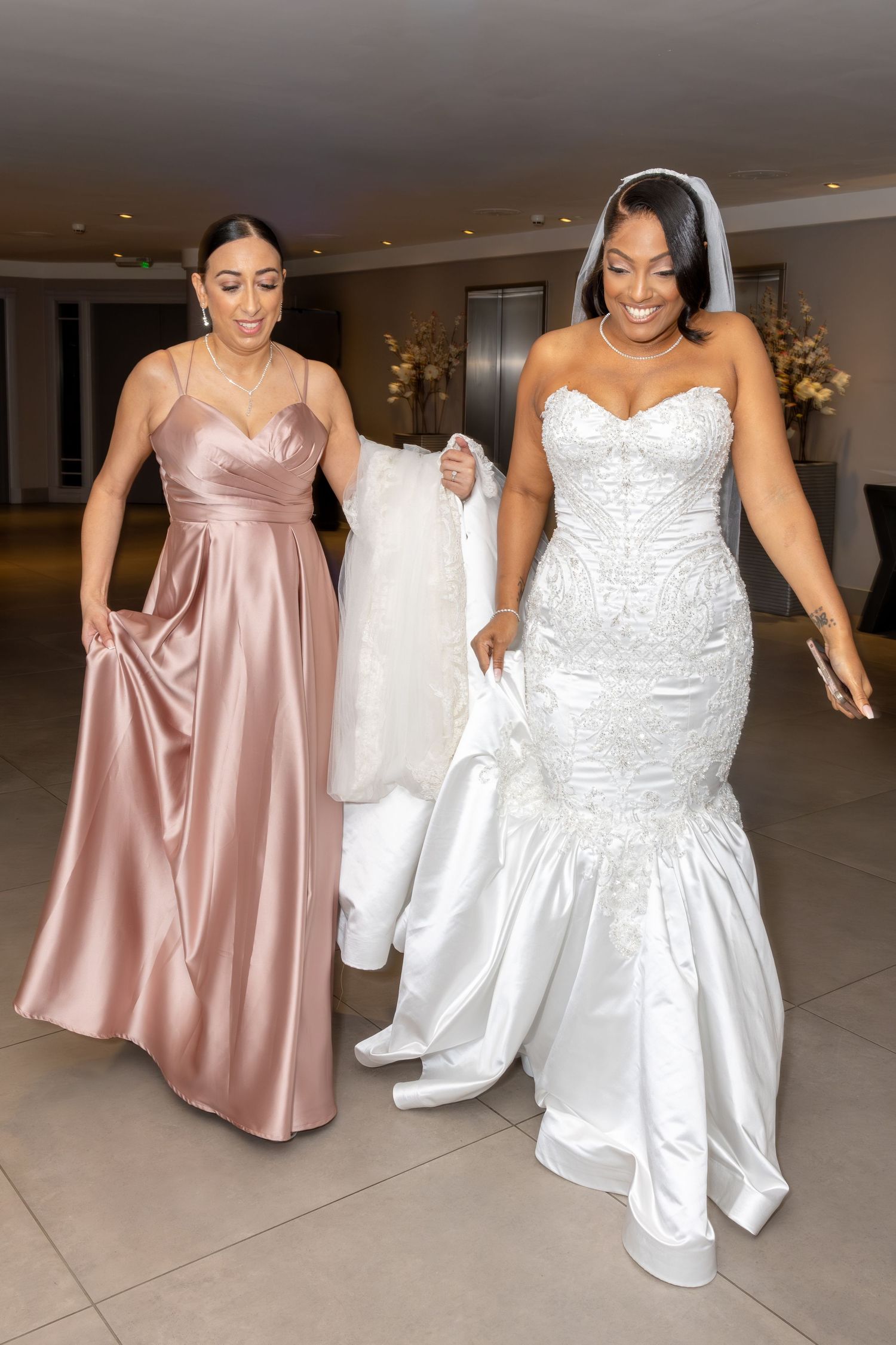 Radiant bride in white mermaid gown walks with bridesmaid in blush pink dress indoors.