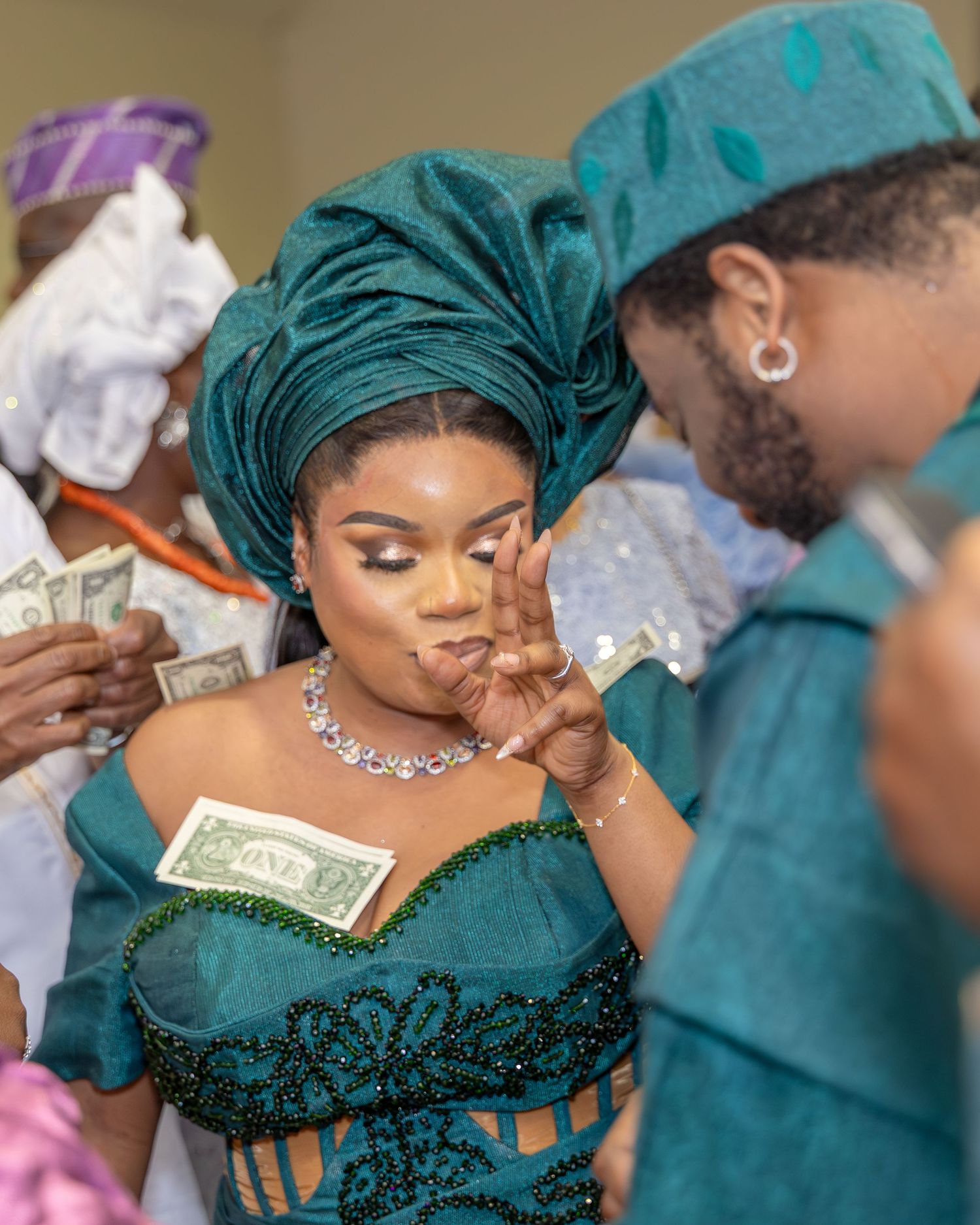 A Nigerian bride in teal aso-oke and groom share a tender moment as money is sprayed during their traditional wedding.