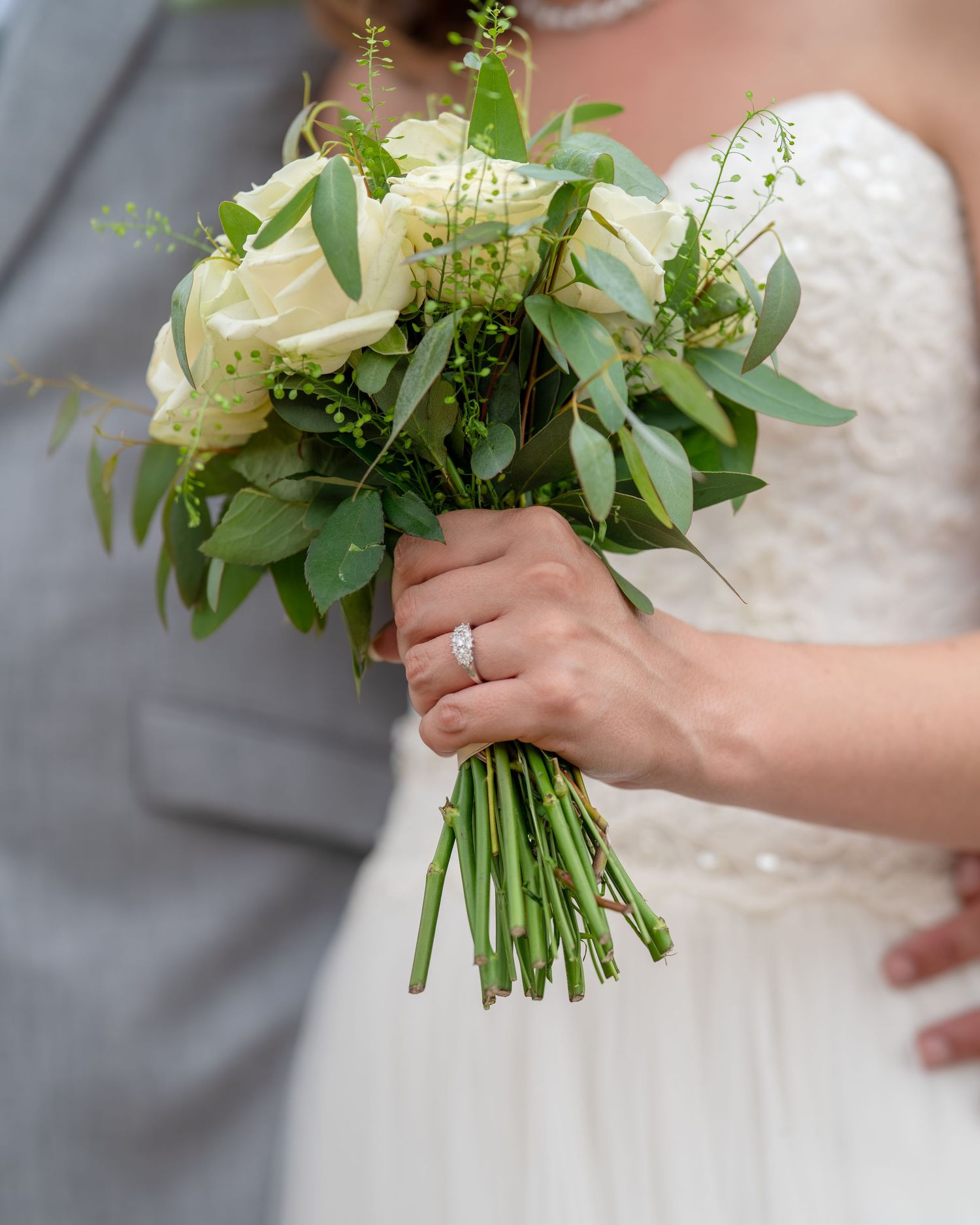 Bride holding a white rose and greenery wedding bouquet with diamond ring visible, groom in gray suit beside her.
