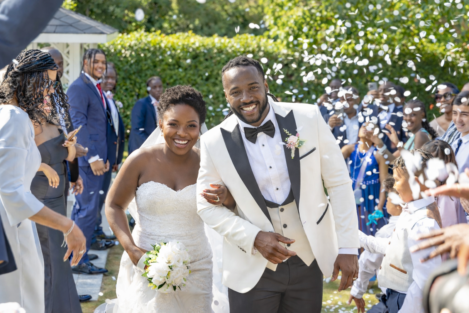 A joyful newlywed couple walks through confetti at their outdoor wedding ceremony surrounded by guests.