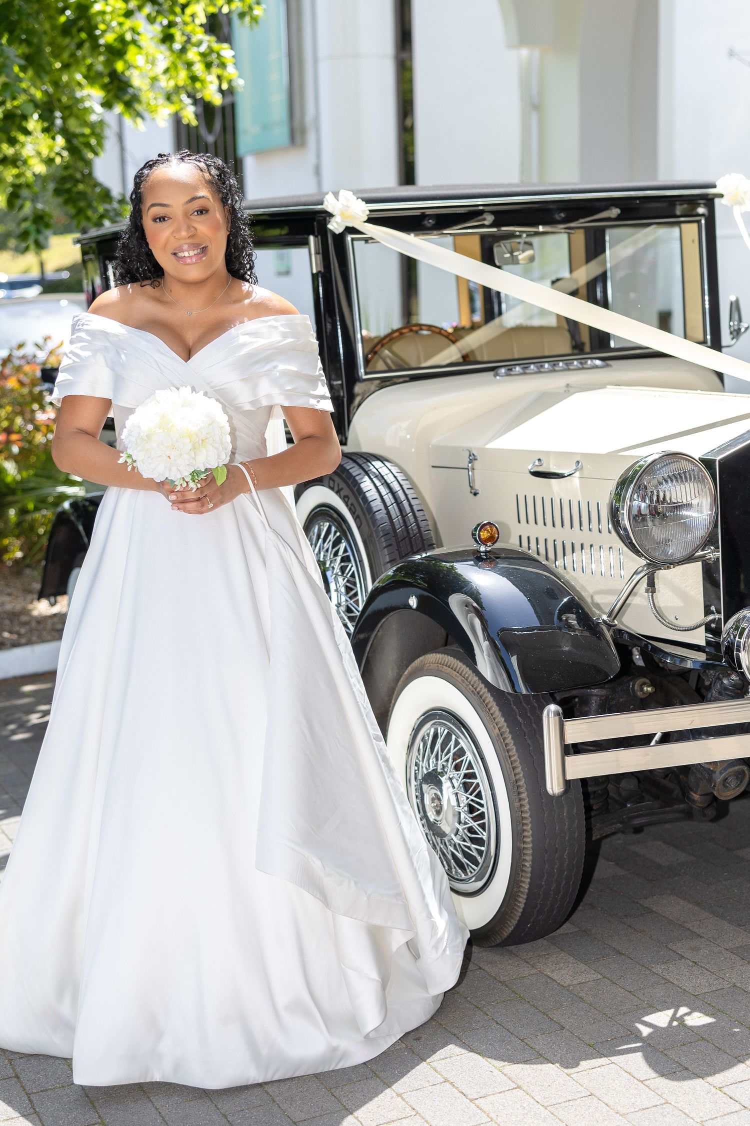 Bride in white off-shoulder gown holds bouquet beside vintage cream wedding car outdoors.