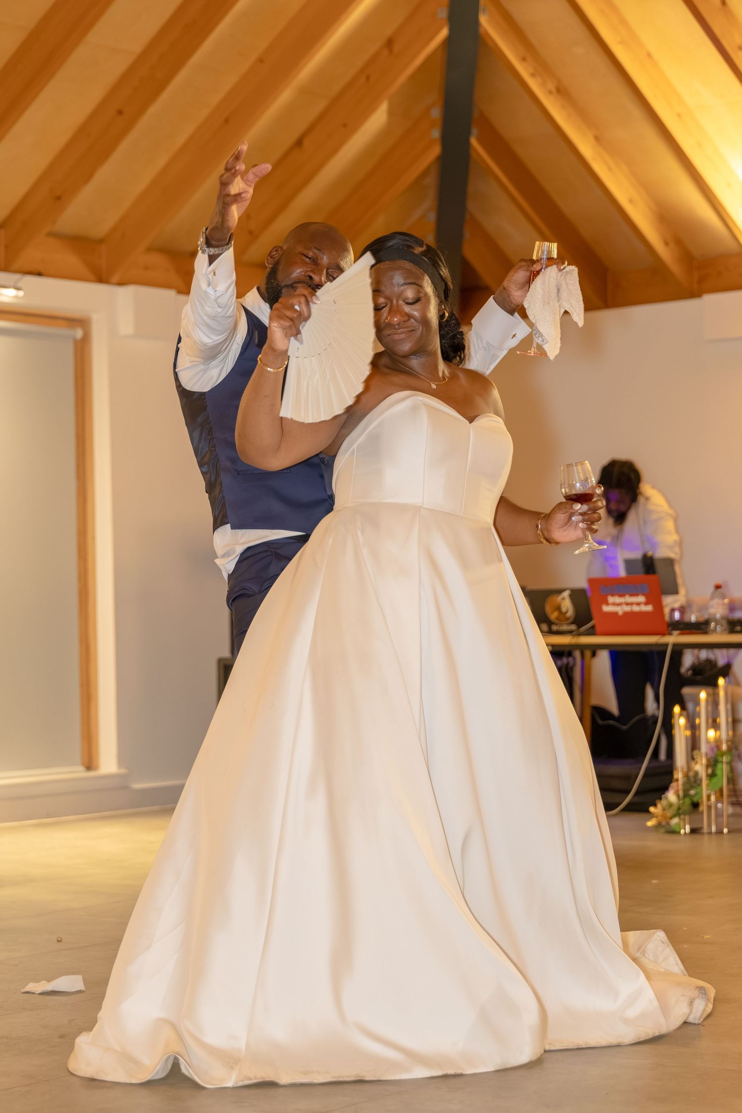 Joyful newlyweds dancing at wedding reception, bride in elegant white ballgown, groom in blue vest, wooden ceiling venue.