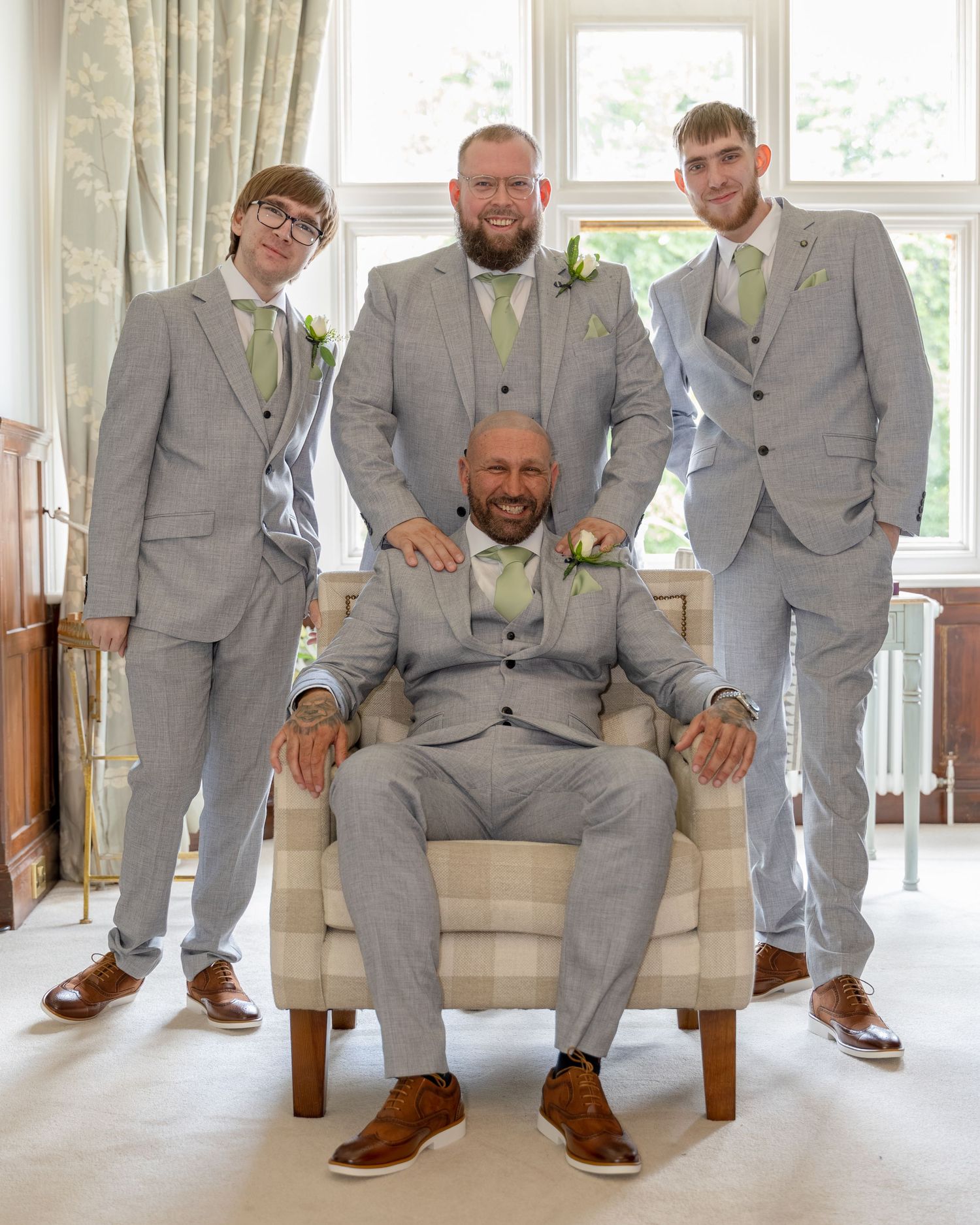 Four men in matching grey suits with green ties pose for a wedding photo, one seated in an armchair.