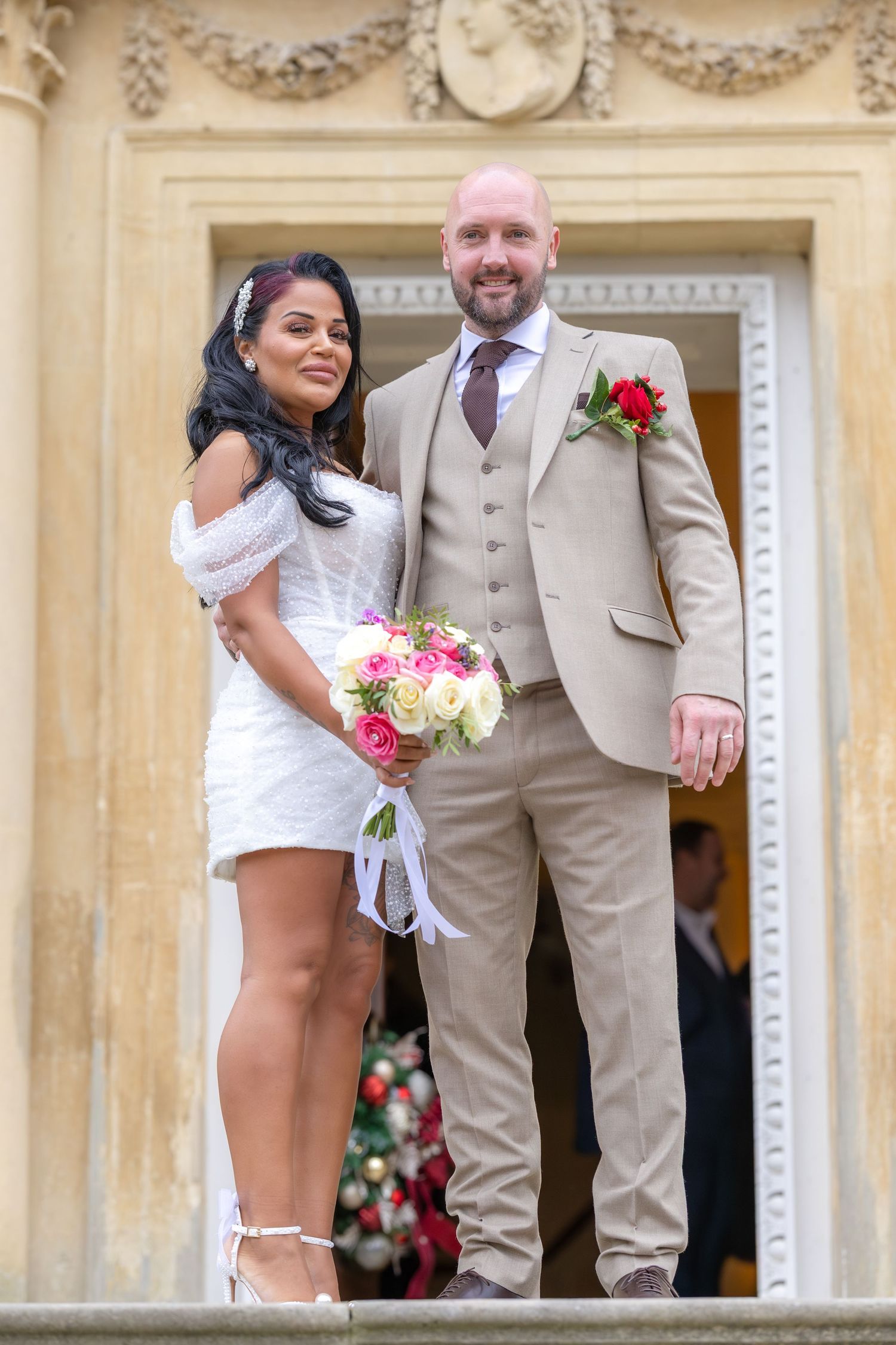 Newlywed couple stand on stone steps, bride in white mini dress holding pink and white bouquet.
