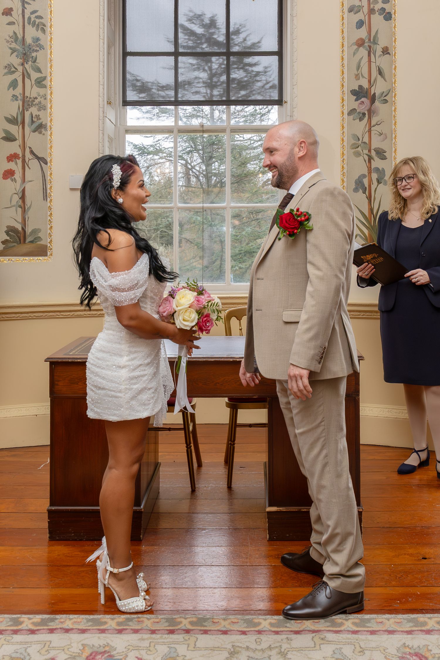 Bride in short white dress holding bouquet facing smiling bald groom in beige suit at wedding ceremony.
