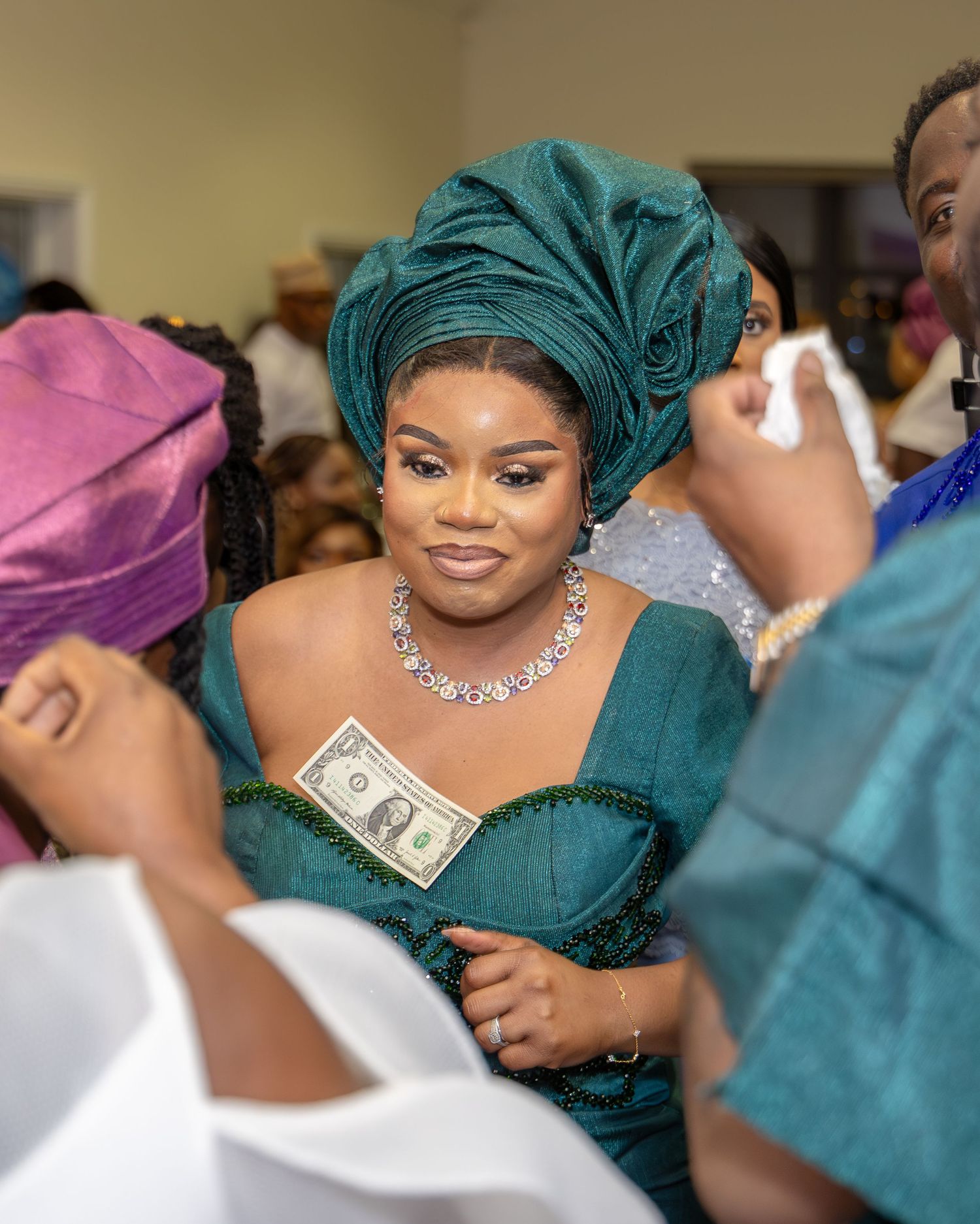 A bride in a teal gele and beaded gown smiles as dollar bills are tucked into her dress during a Nigerian wedding.