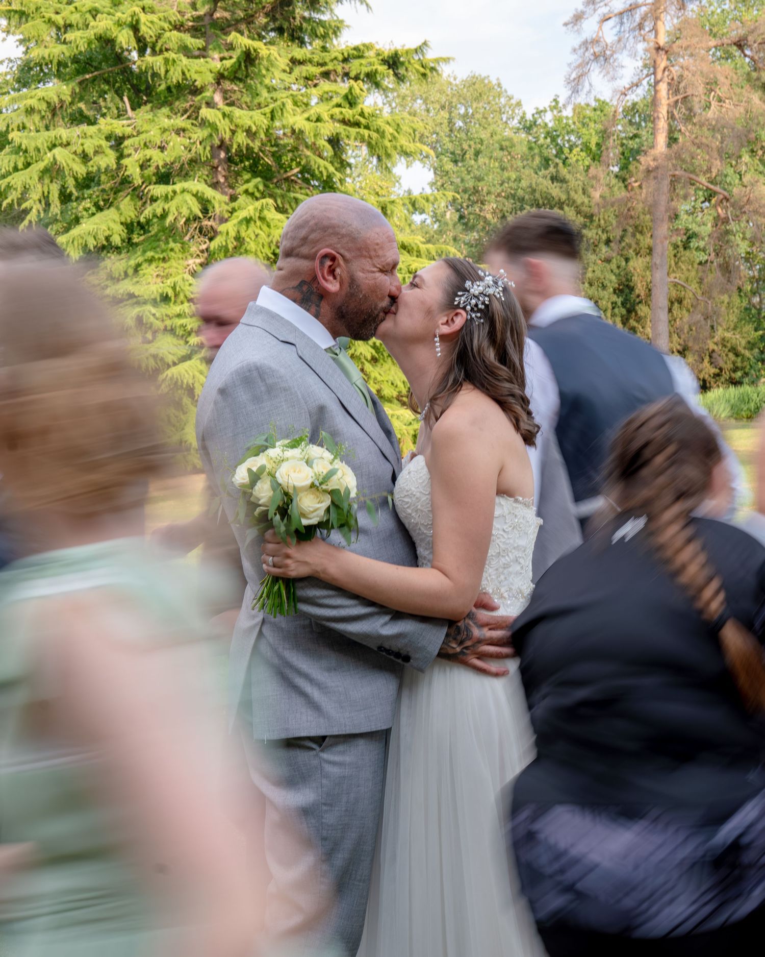 Bride and groom share a kiss surrounded by wedding guests outdoors, bride holds white bouquet, groom wears gray suit.