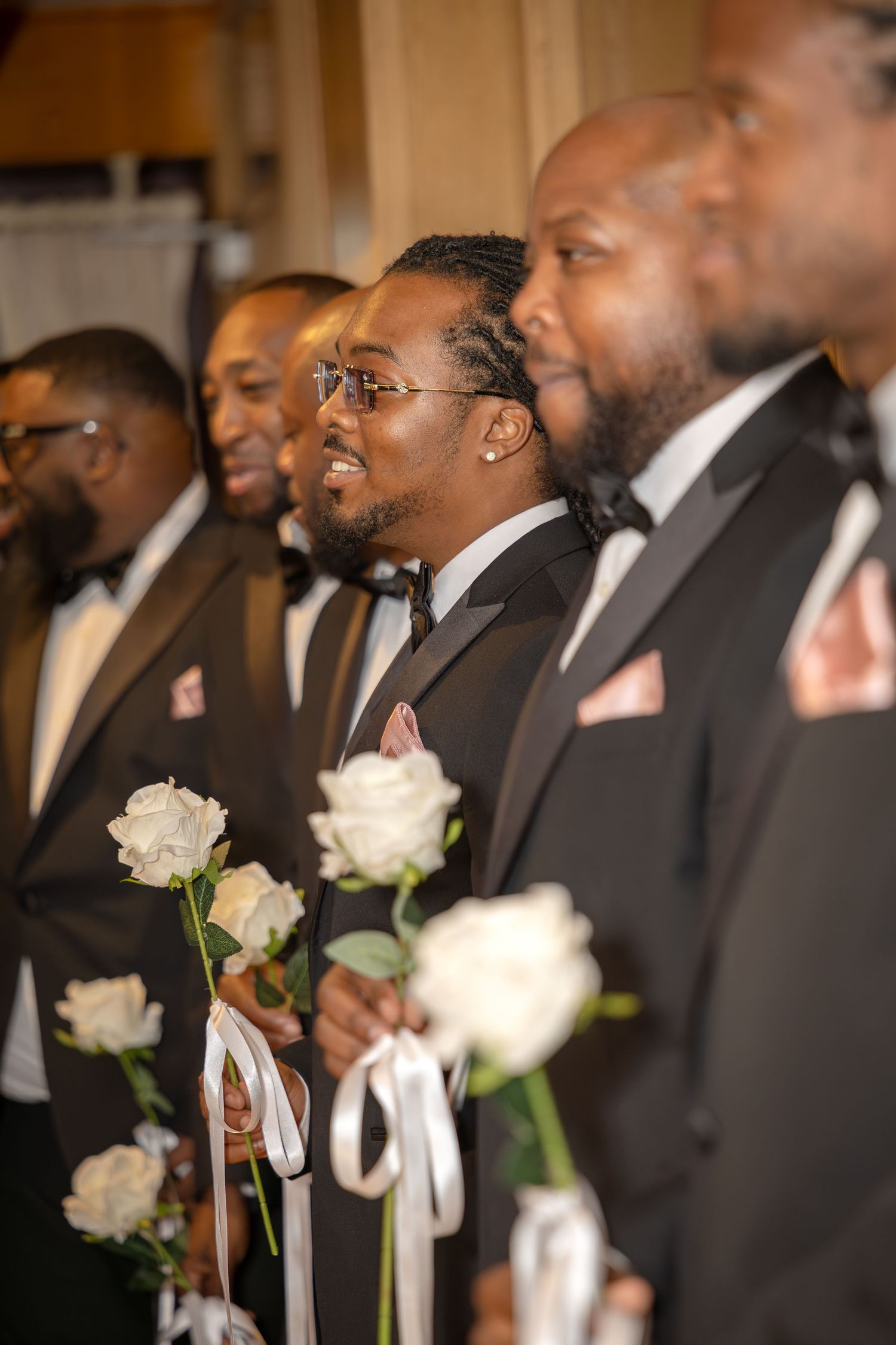 Groomsmen in black tuxedos holding white roses line up during a wedding ceremony processional.