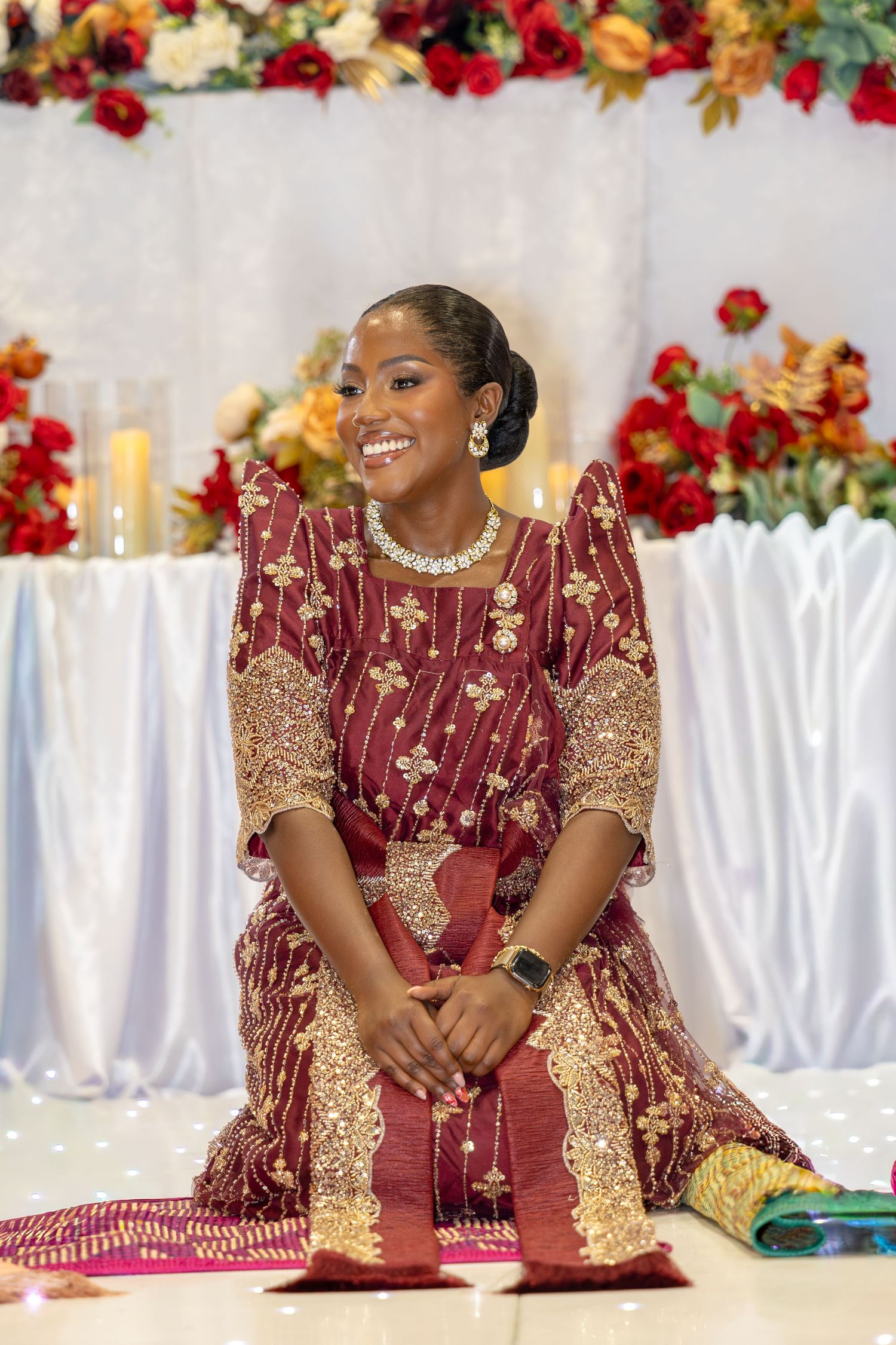 Radiant bride in embroidered burgundy gold African traditional gown kneels smiling before a floral decorated altar.