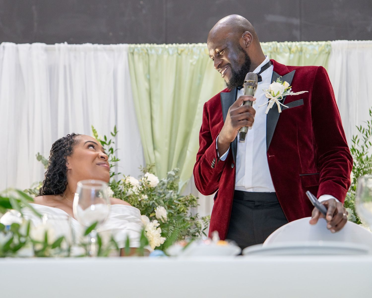 Groom in red velvet tuxedo gives wedding speech to smiling bride at floral-decorated reception table.