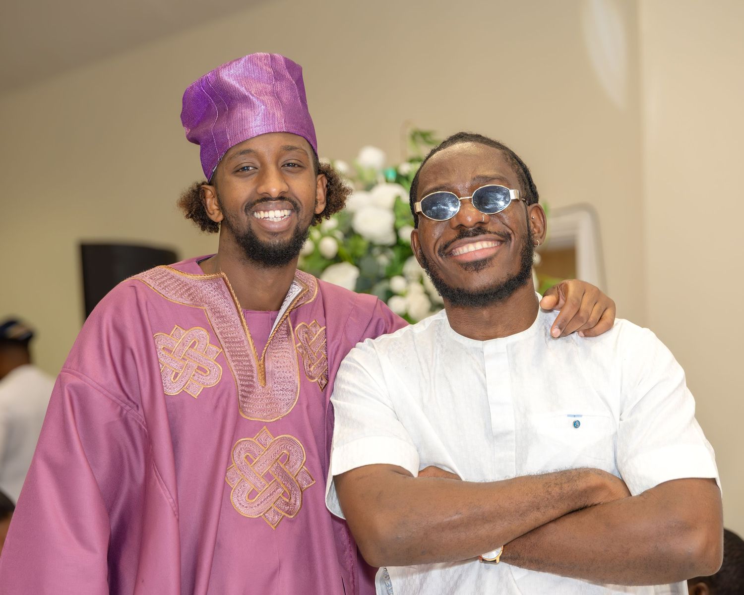Two men smiling at Nigerian traditional wedding, one wearing purple agbada with matching fila cap, other in white outfit.