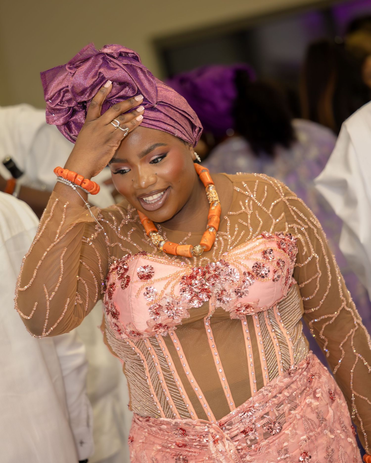 Woman in pink beaded dress and purple headwrap smiles at a cultural celebration event.