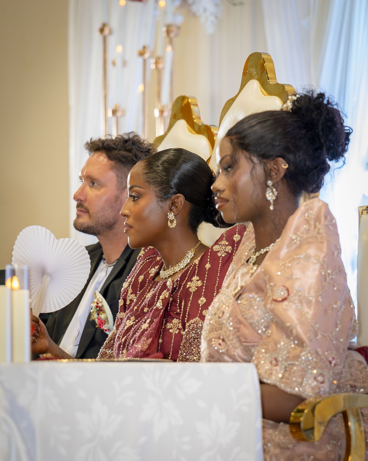 Interracial couple and bridesmaid seated at wedding ceremony altar with candles and gold decor.