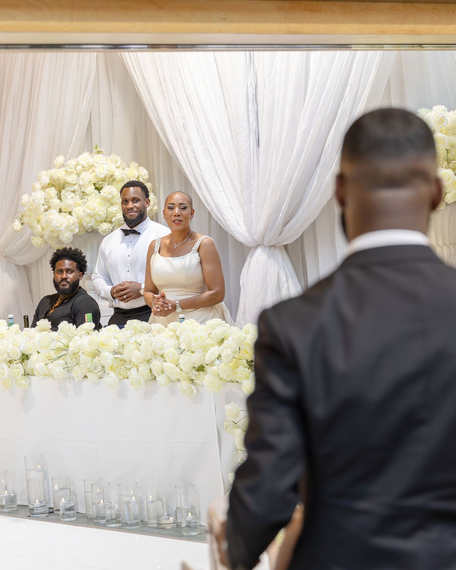 Elegant wedding reception with bride and groom at floral table, white drapes and roses backdrop.