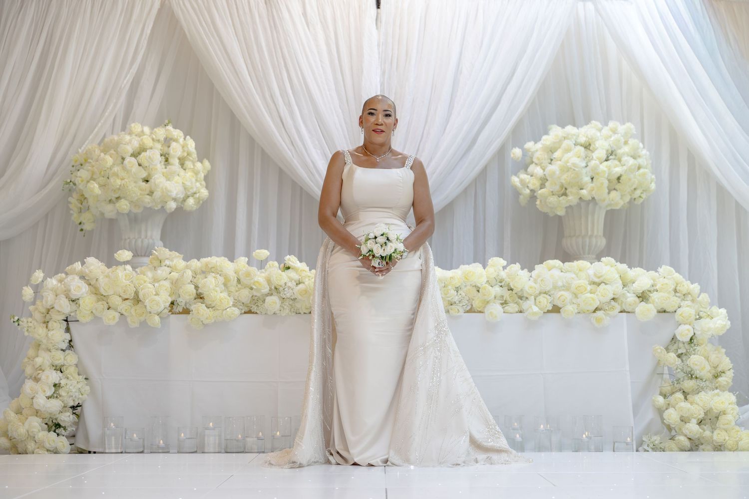 Bride in elegant white gown holding bouquet stands before floral arrangements and draped backdrop.