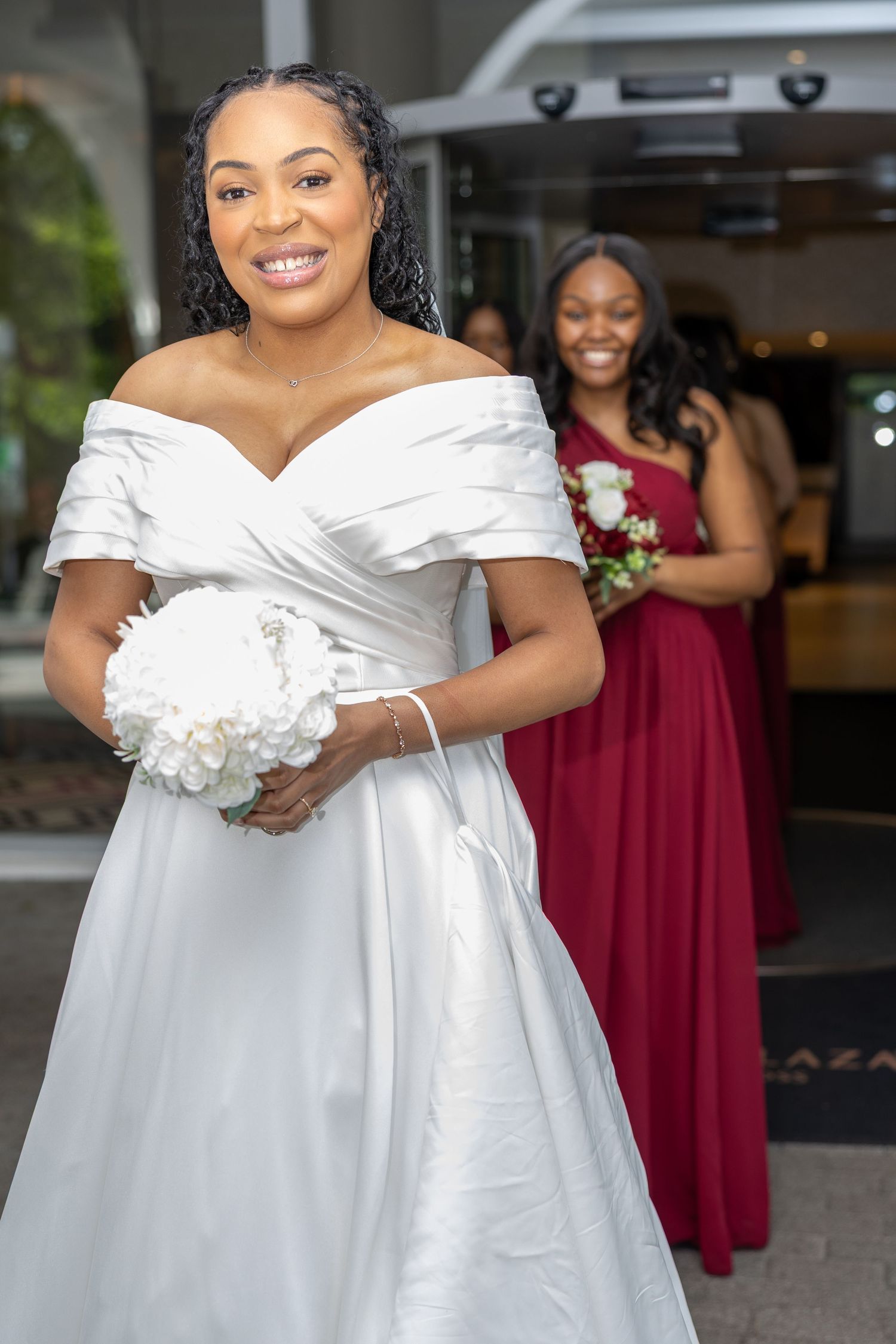 Smiling bride in white off-shoulder gown holds bouquet, bridesmaid in red dress stands behind her.