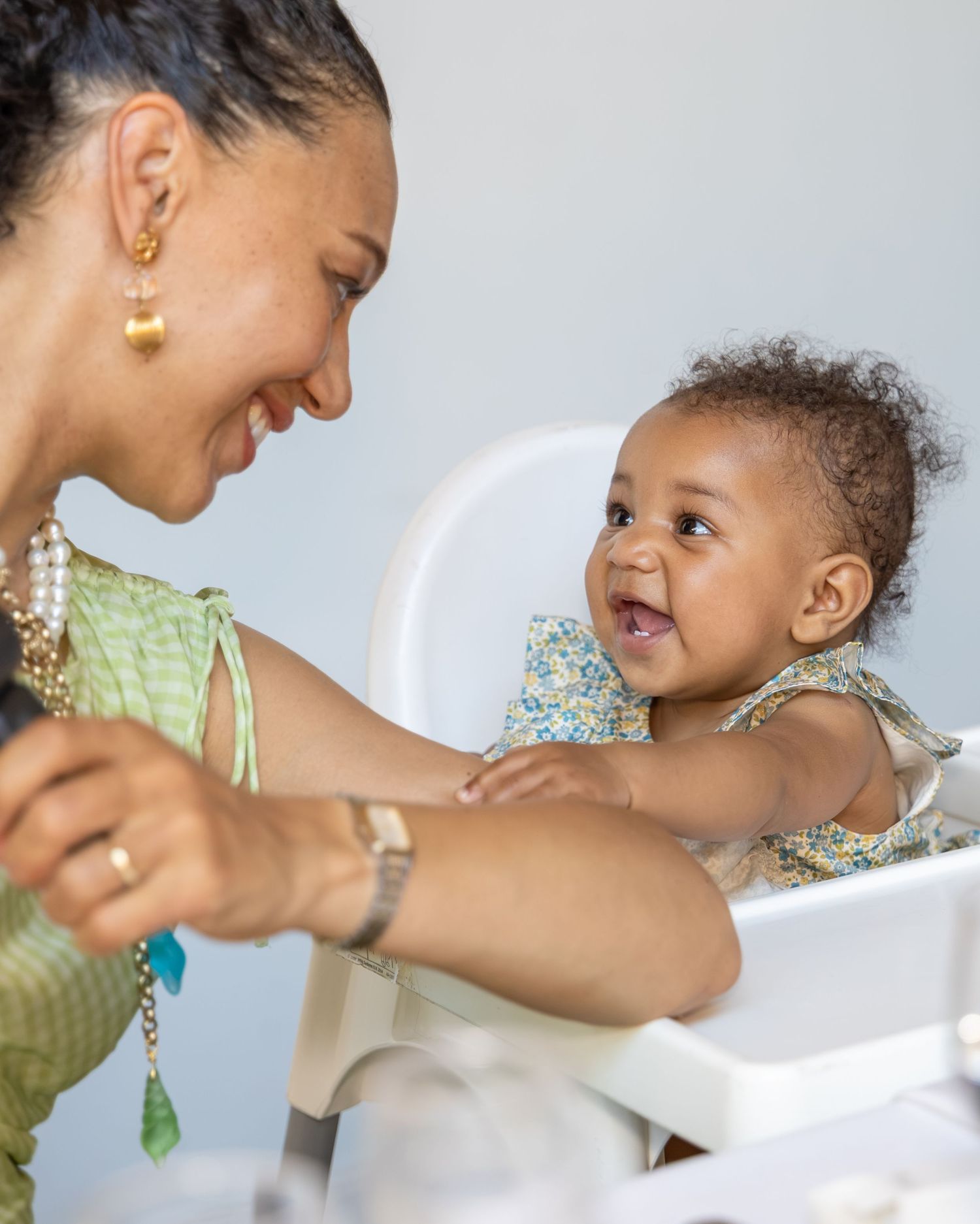 A smiling woman in a green top interacts joyfully with a laughing baby in a floral outfit seated in a high chair.